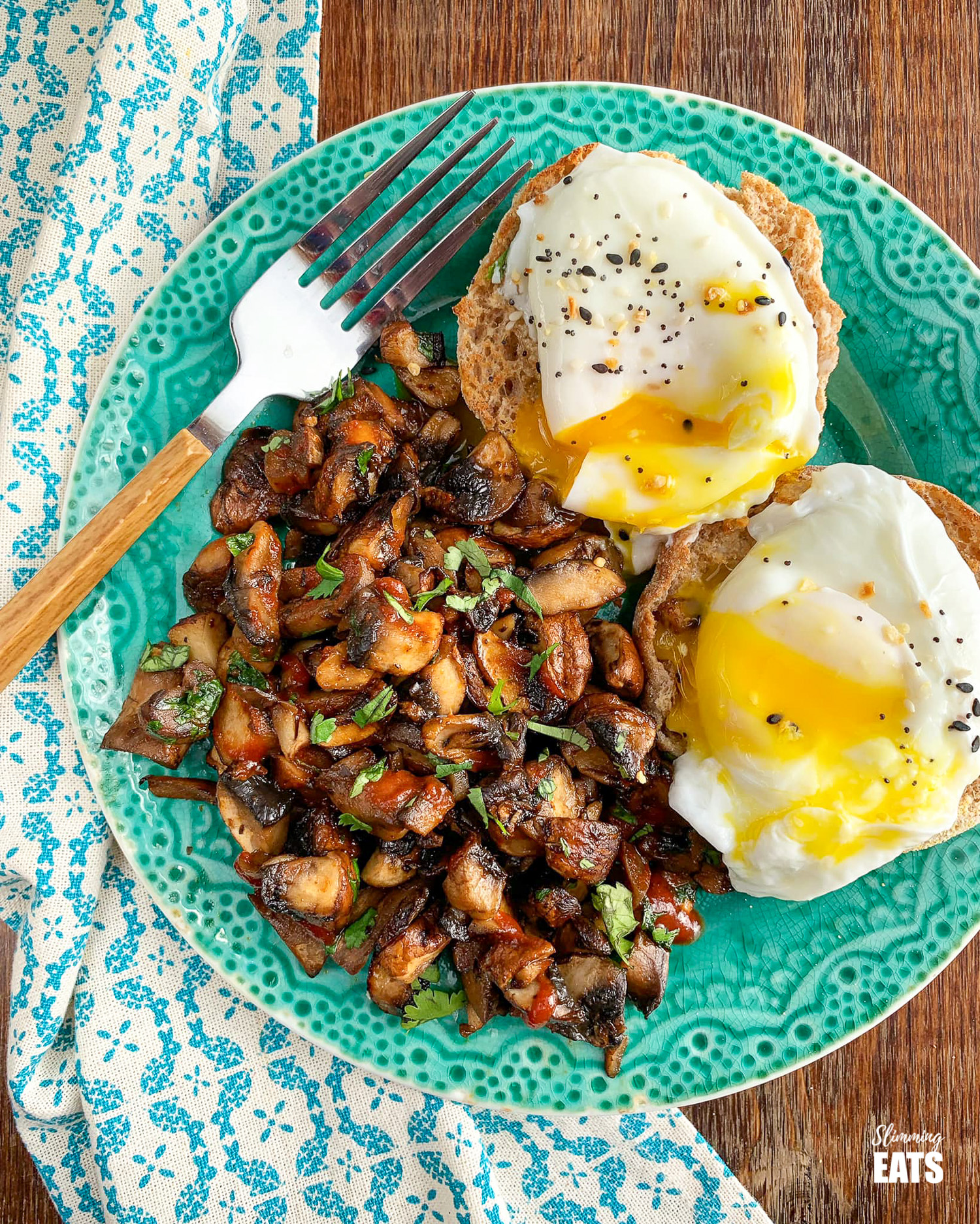 toasted wholewheat muffin topped with poached eggs on the side or sriracha style mushrooms on a teal plate with fork to the left.