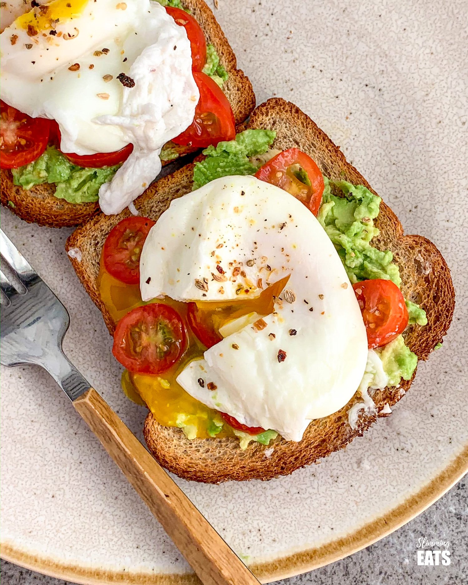 poached eggs over wholewheat toast with mashed avocado and sliced cherry tomatoes on beige rimmed cream plate with fork to the let