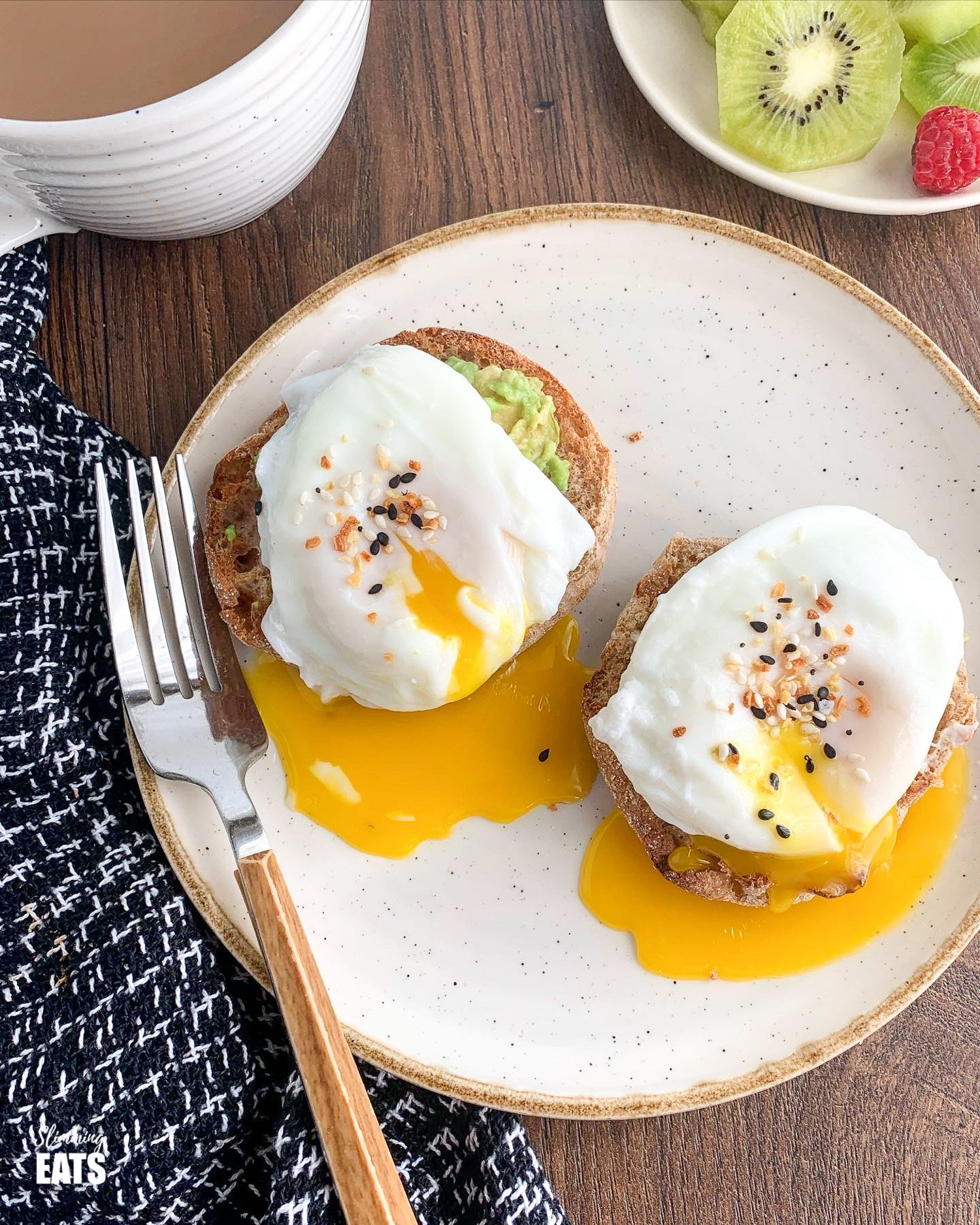 poached eggs over mashed avocado on toasted English muffin on white plate with yellow rim with mug of coffee and bowl of mixed fruit