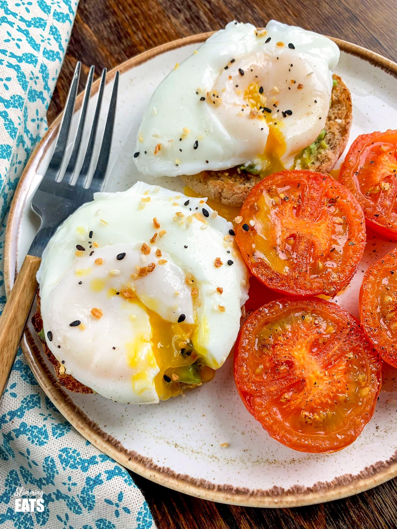 poached eggs over toasted wholewheat muffins with mashed avocado and grilled tomatoes on brown rimmed cream plate