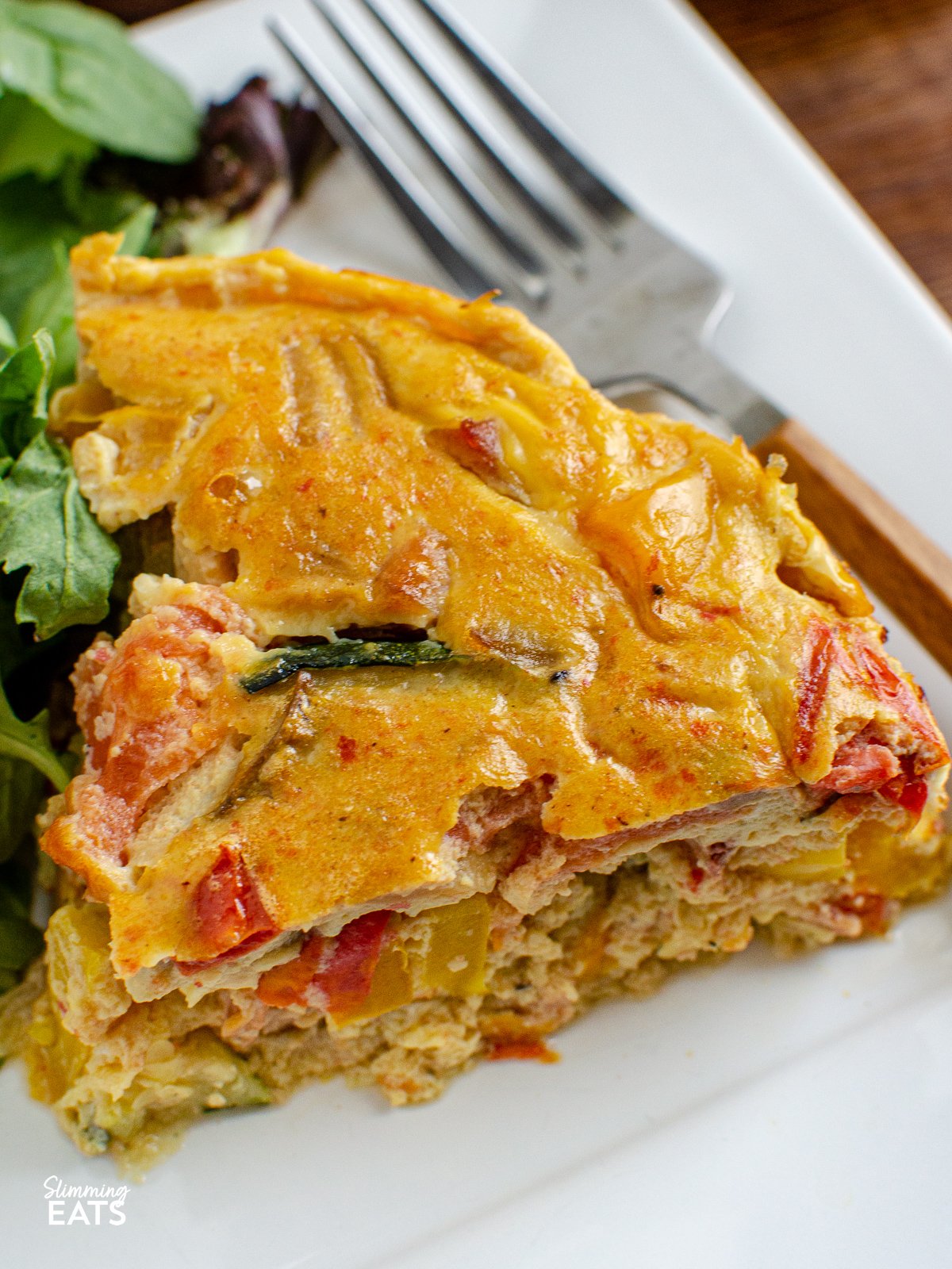 Enjoy a delicious and healthy alternative to the classic quiche. This dairy free crustless bacon vegetable quiche is packed with protein and flavourful vegetables and is perfect for those who need to avoid dairy. close up of Dairy Free Crustless Bacon and Vegetable Quiche on white plate with salad