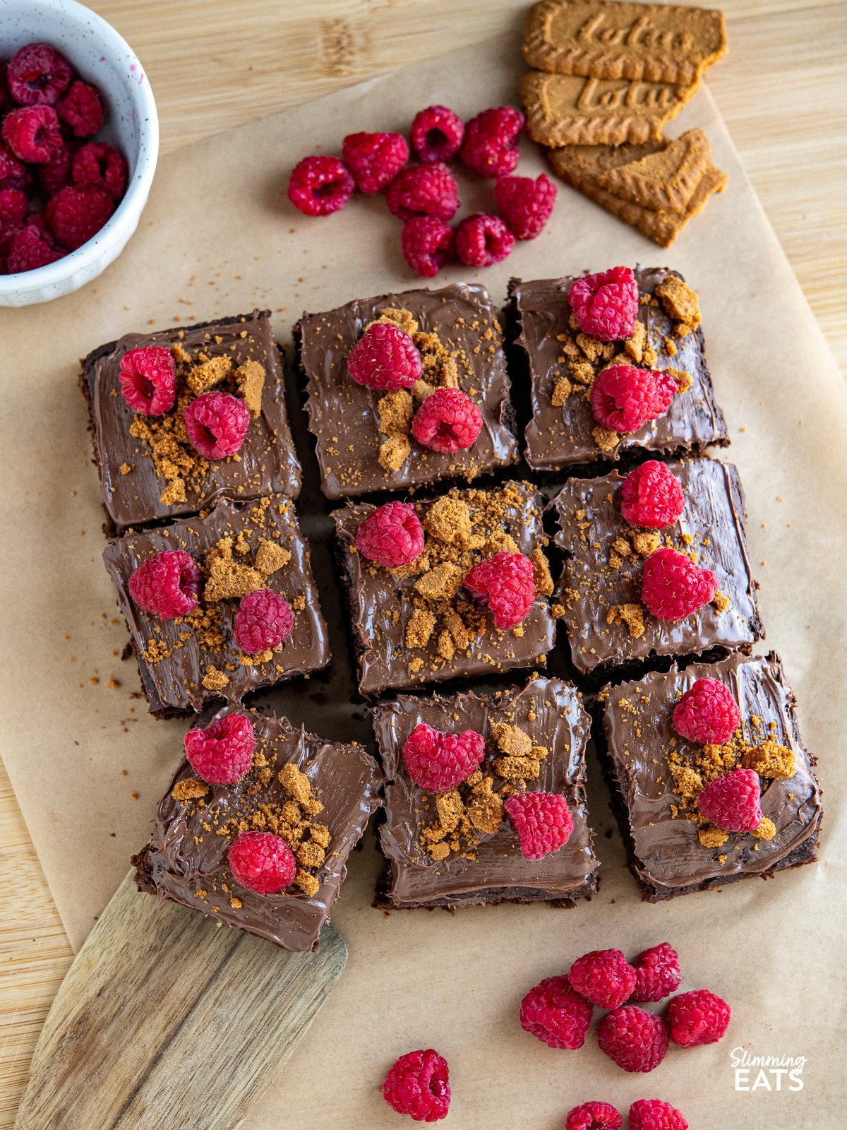 Love a slice of guilt-free cake? Check out this delicious low calorie chocolate cake for only 158 calories a slice, topped with nutella spread, crumbled lotus biscoff cookies and fresh raspberries. sliced Raspberry Nutella Chocolate Cake on a wooden board with scattered raspberries and lotus cookies