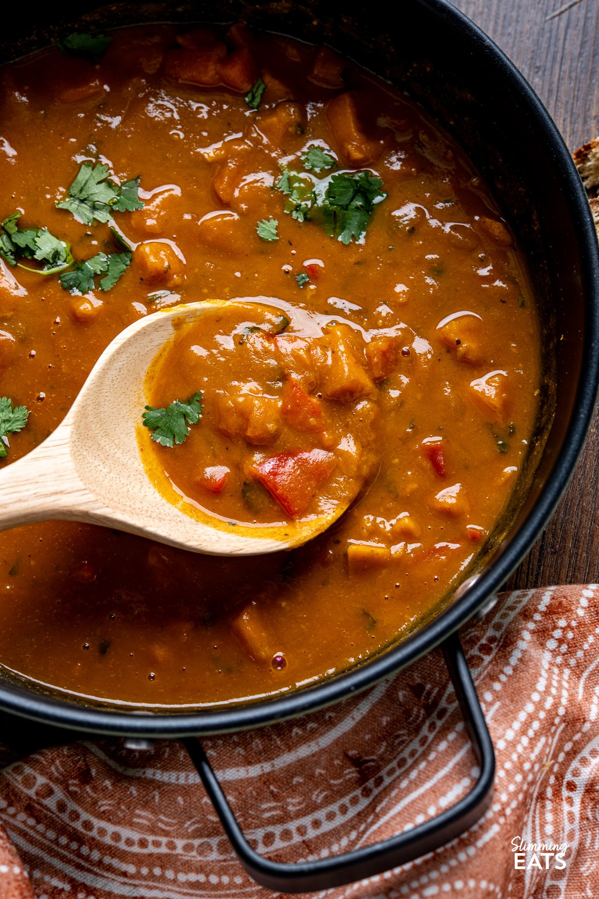 
close up of Sweet Potato, Red Pepper and Carrot Soup in a black double handled pan with wooden spoon