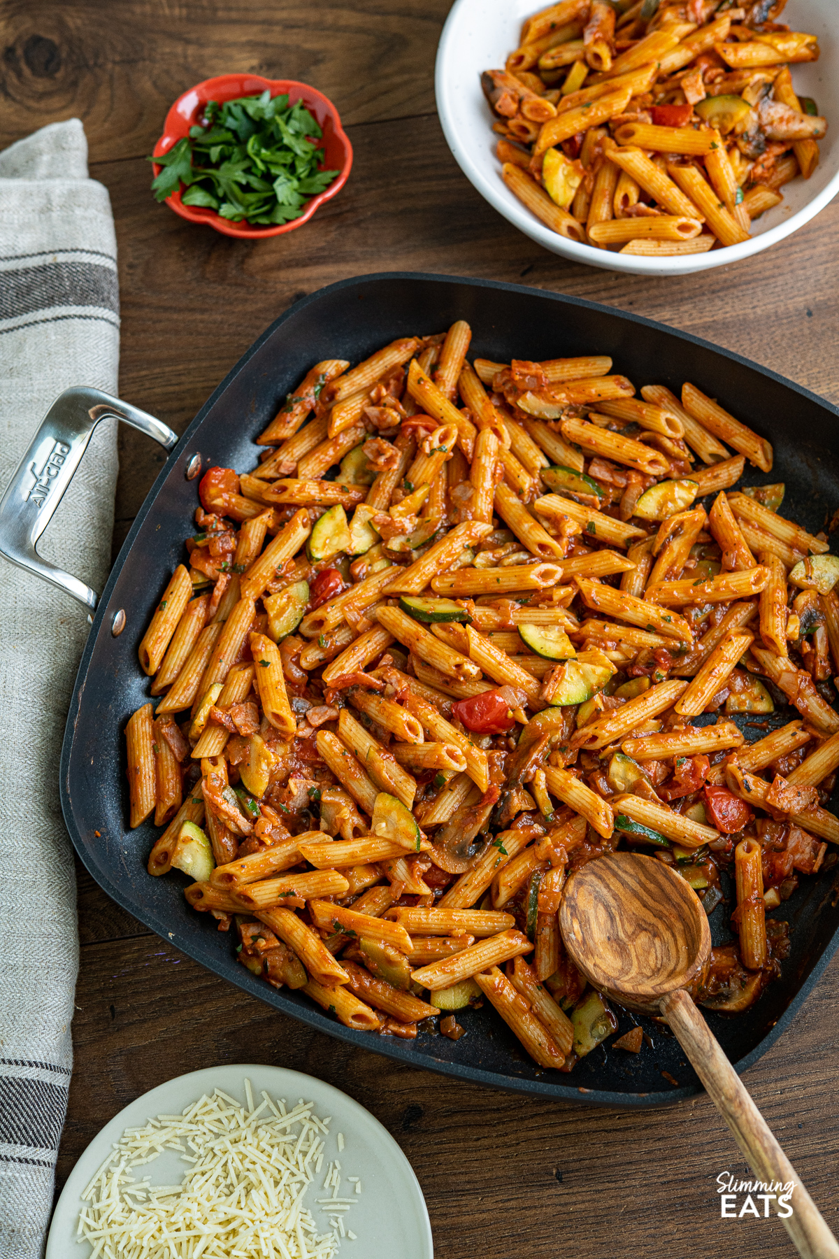Mushroom, Bacon, Tomato and Zucchini Pasta in square all-clad skillet with bowl of pasta and small terracotta pinch bowl of parsley in background