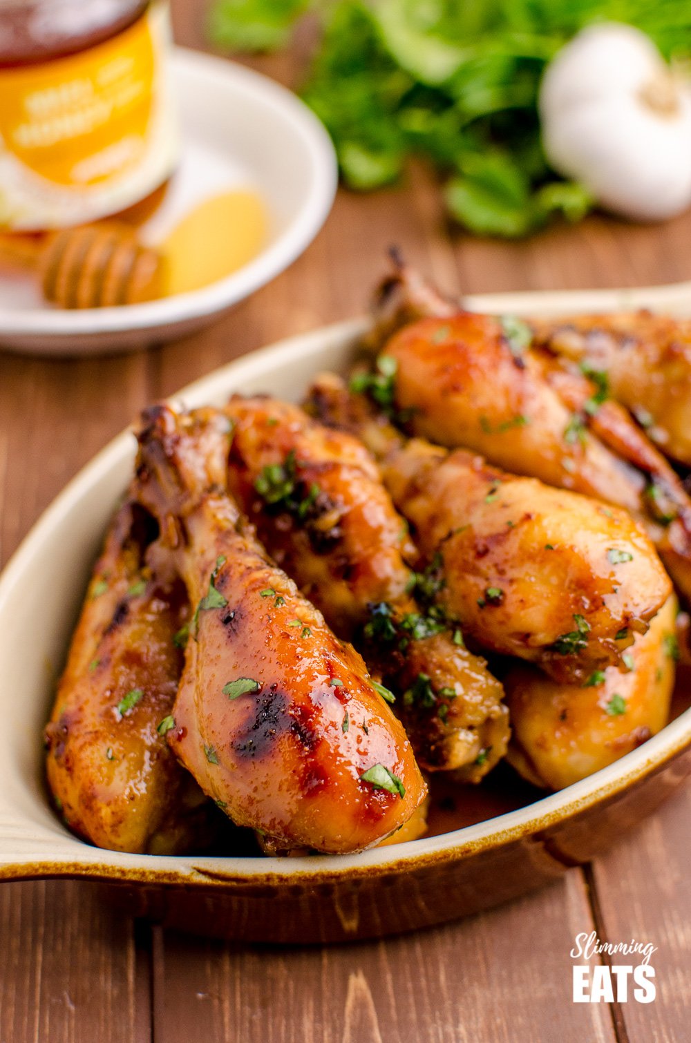 Honey and Garlic Chicken Drumsticks in dish with honey, garlic and herbs in background