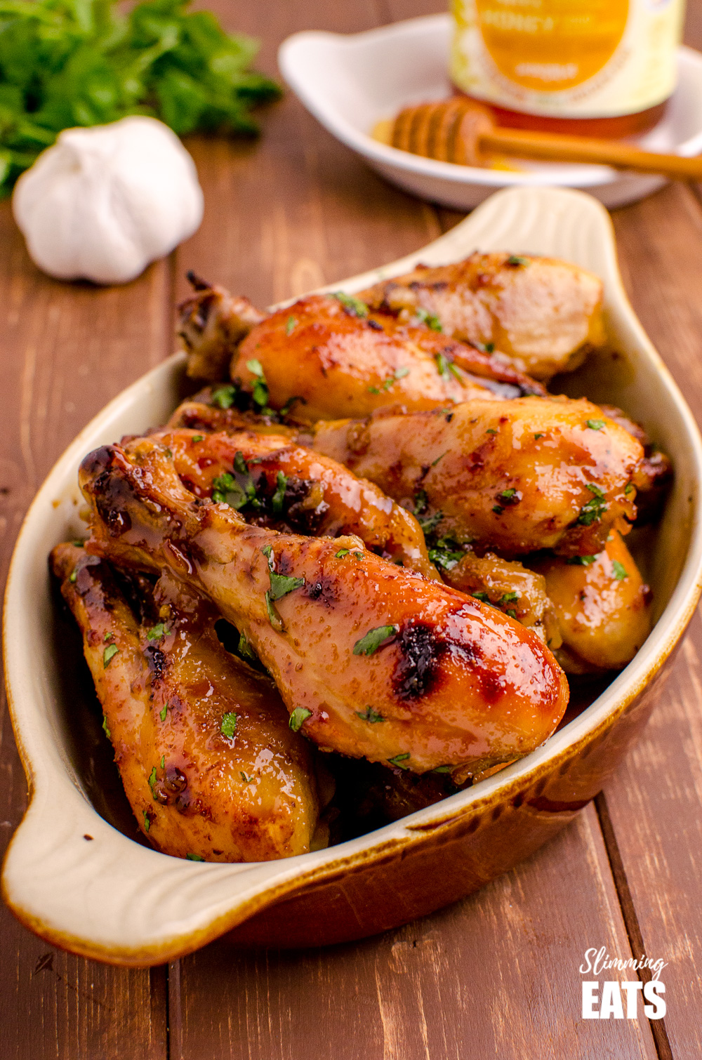 Honey and Garlic Chicken Drumsticks in dish on wooden board with honey, garlic and herbs in background