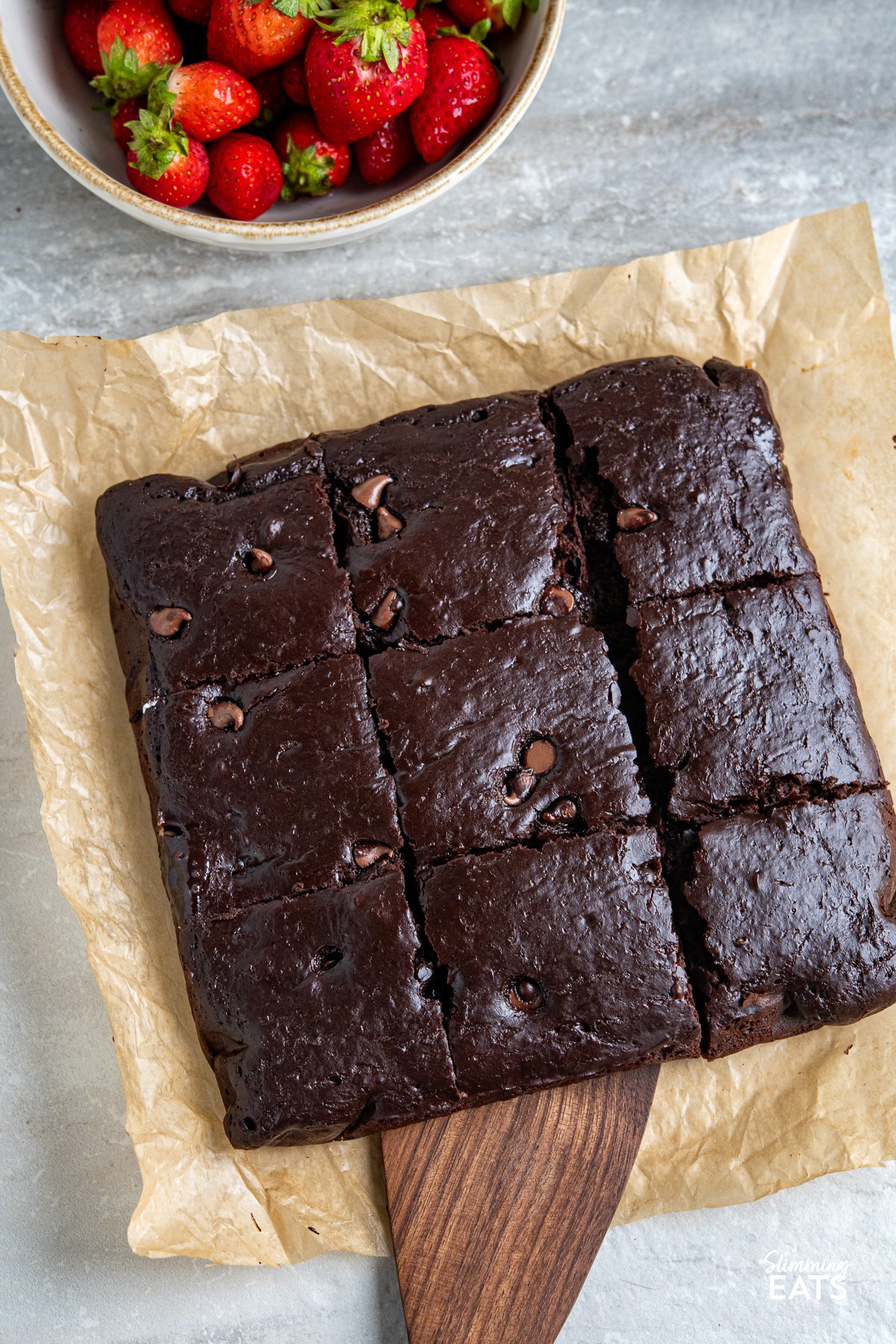 Squidgy chocolate cake cut into 9 squares on parchment paper, with one slice being lifted by a spatula. A bowl of fresh strawberries sits in the background.