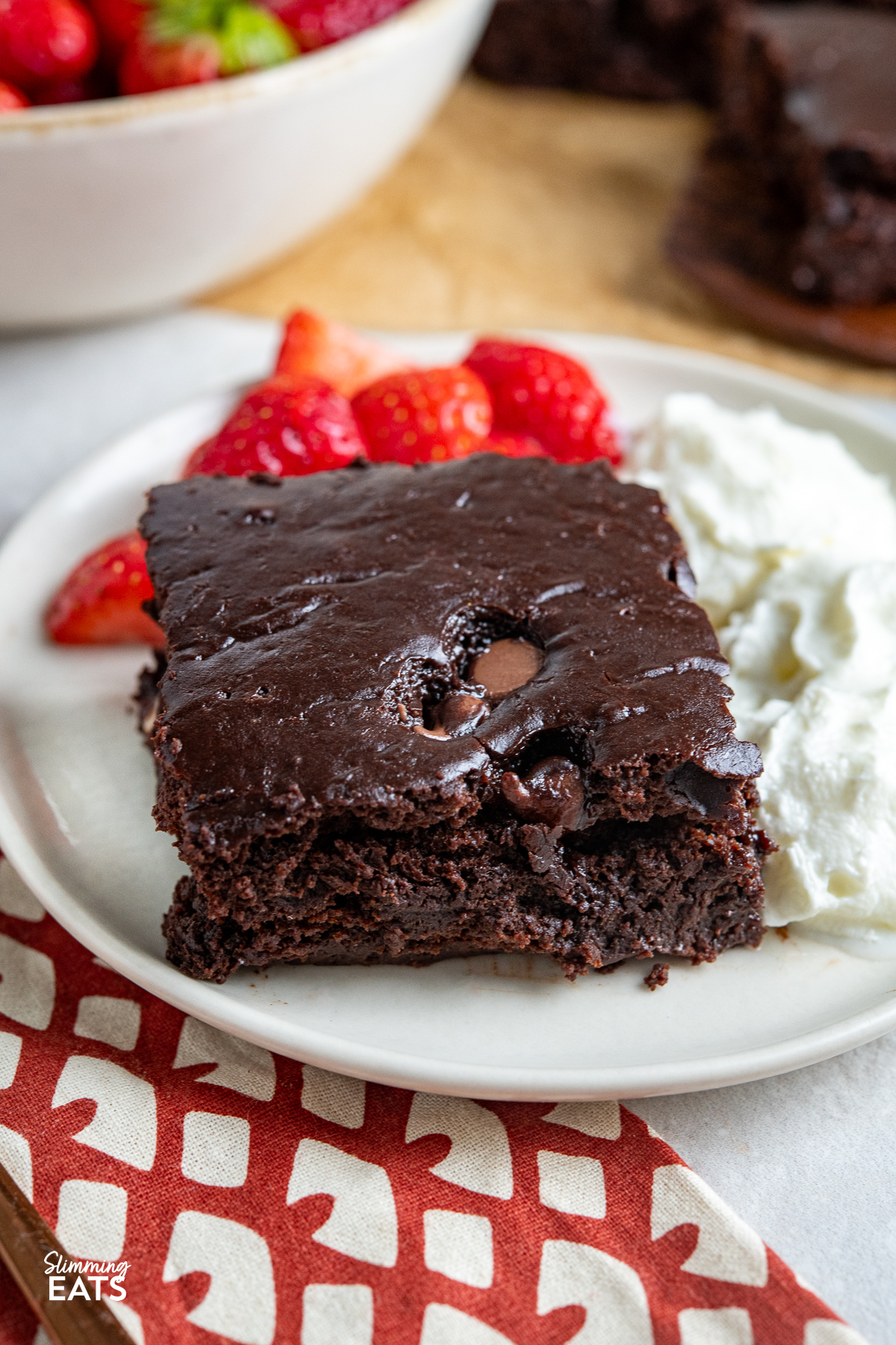 Slice of squidgy chocolate cake on a cream-colored plate, accompanied by aerosol cream and strawberries, with more cake and a bowl of strawberries visible in the background.