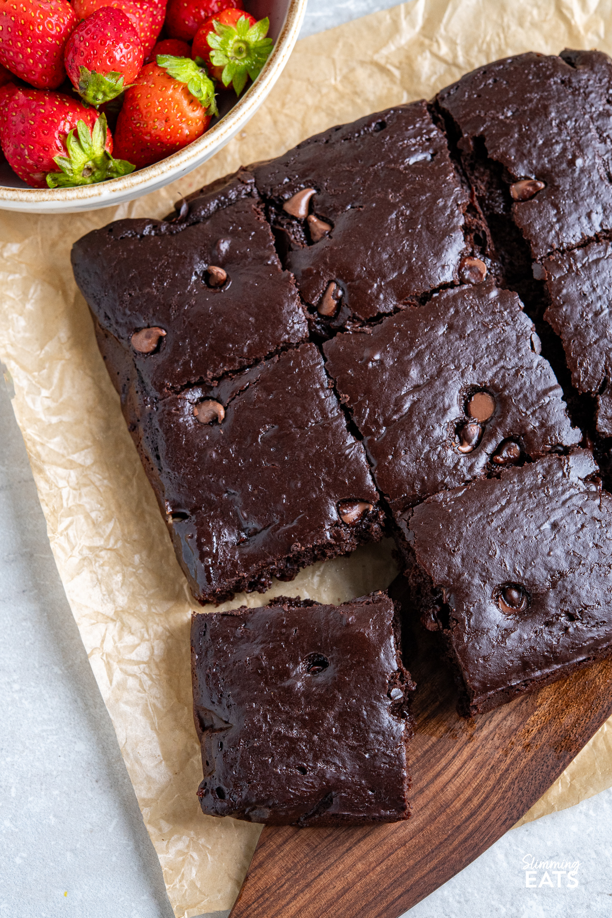 Squidgy chocolate cake cut into 9 squares on parchment paper, with one slice being lifted by a spatula. A bowl of fresh strawberries sits in the background.
