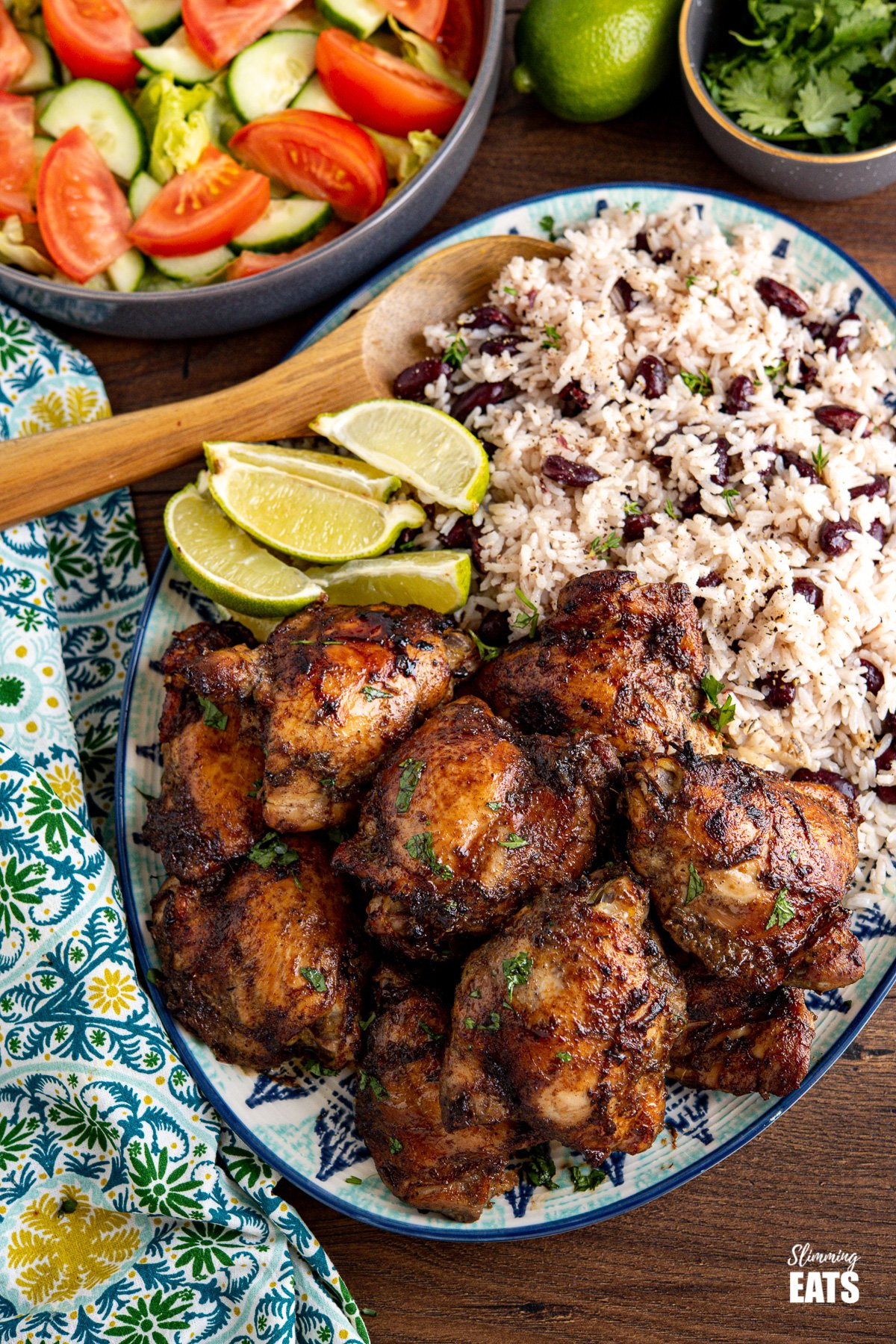 large patterned oval plate with Jamaican Jerk Chicken with Rice and Peas and lime wedges, salad, limes and small grey bowl of cilantro in the background