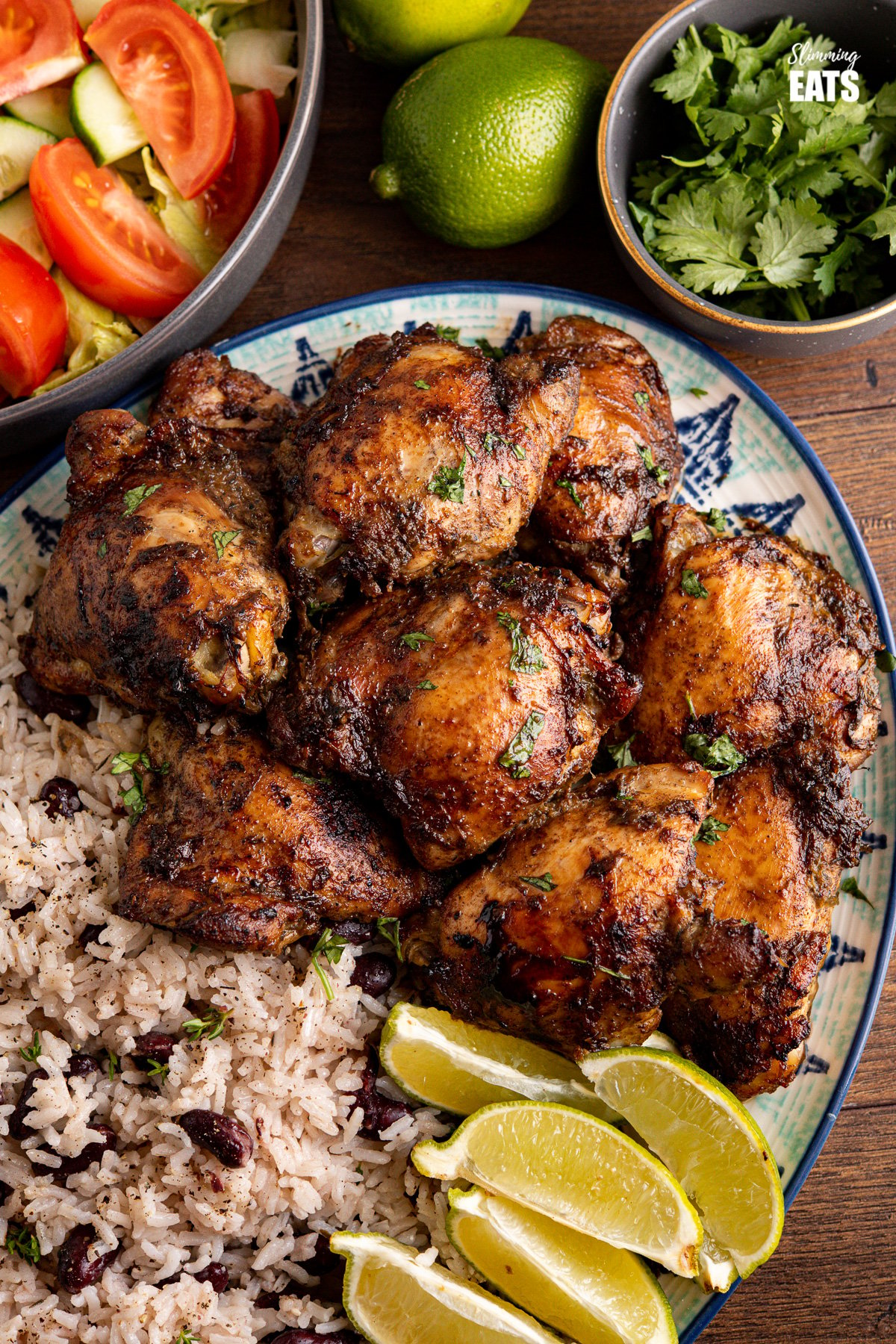 close up of large patterned oval plate with Jamaican Jerk Chicken with Rice and Peas and lime wedges, salad, limes and small grey bowl of cilantro in the background