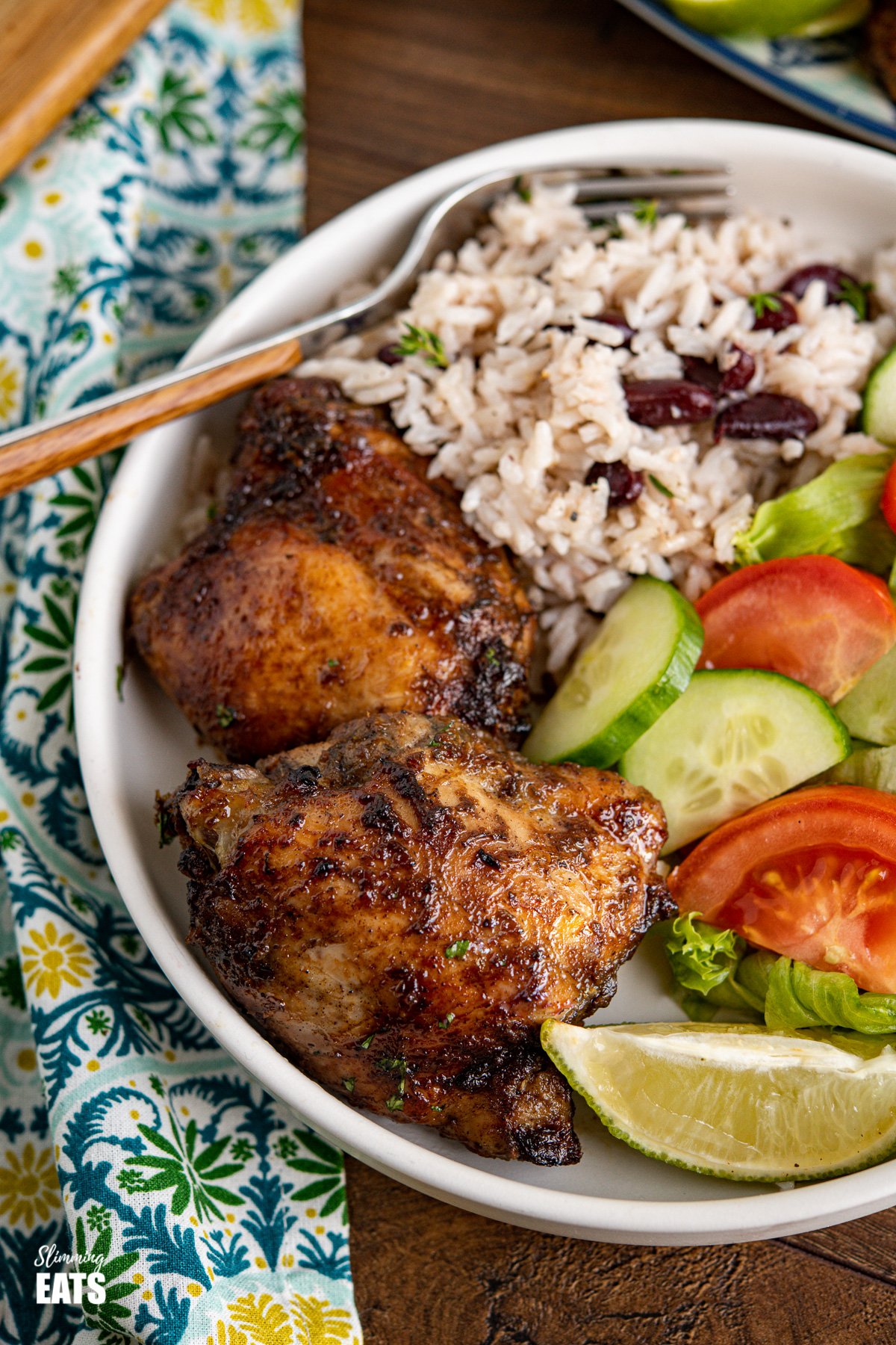 close up of Jamaican Jerk Chicken with Rice and Peas in a white bowl with a lime wedge and salad