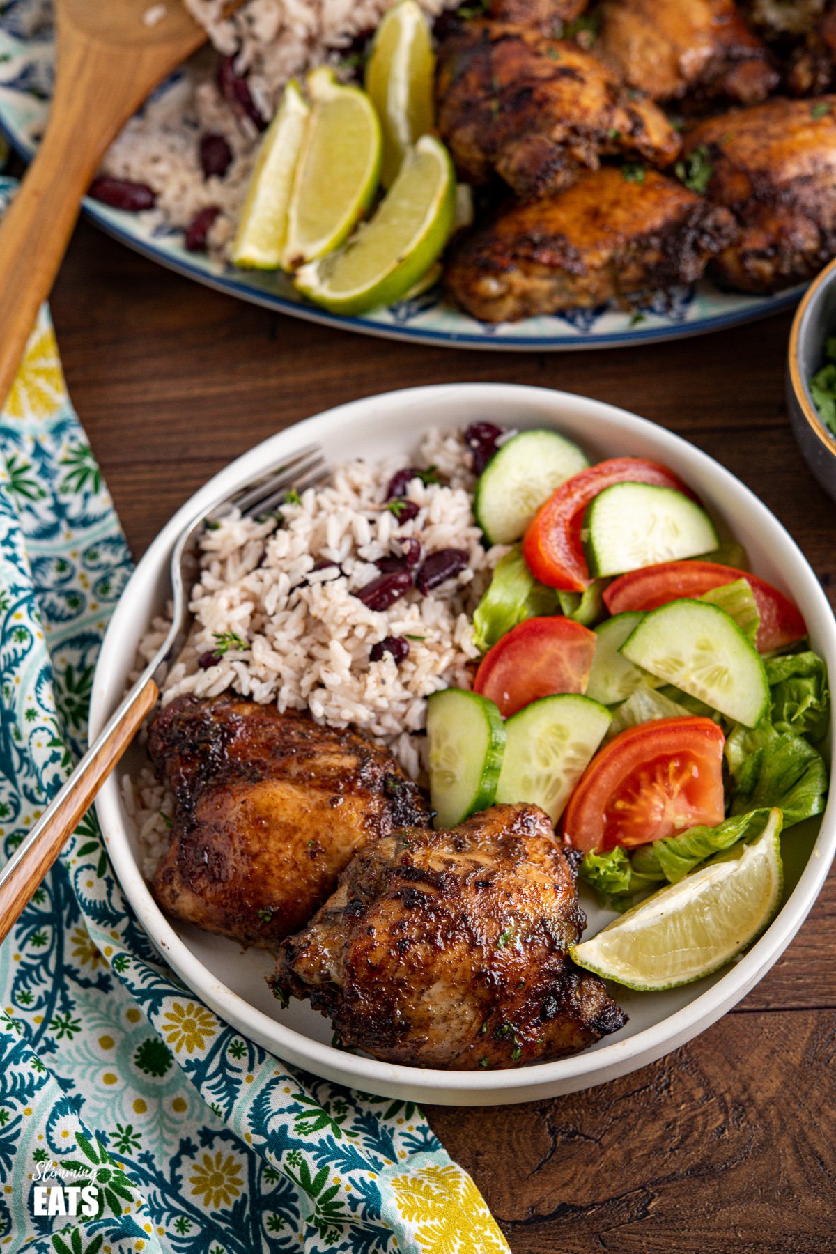 Jamaican Jerk Chicken with Rice and Peas in a white bowl with a lime wedge and salad, oval platter of jerk chicken and rice and peas in background