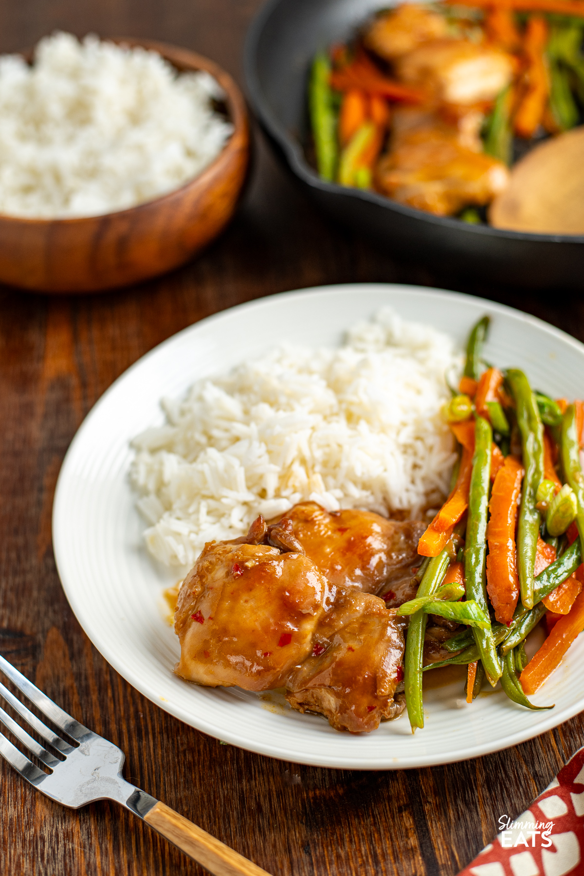 Tender chicken thighs, crisp green beans, and vibrant carrots coated in a delectable Thai sweet chili sauce, presented on a clean white plate next to a mound of aromatic jasmine rice. A silver fork rests to the left, eagerly anticipating the culinary adventure ahead.