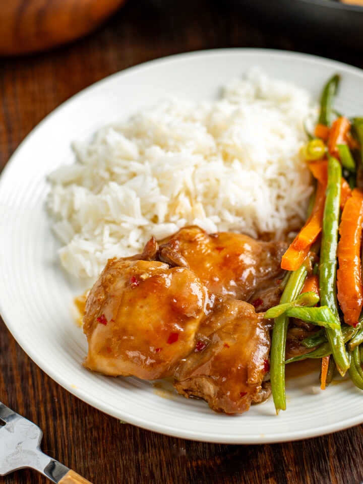 Tender chicken thighs, crisp green beans, and vibrant carrots coated in a delectable Thai sweet chili sauce, presented on a clean white plate next to a mound of aromatic jasmine rice. A silver fork rests to the left, eagerly anticipating the culinary adventure ahead.