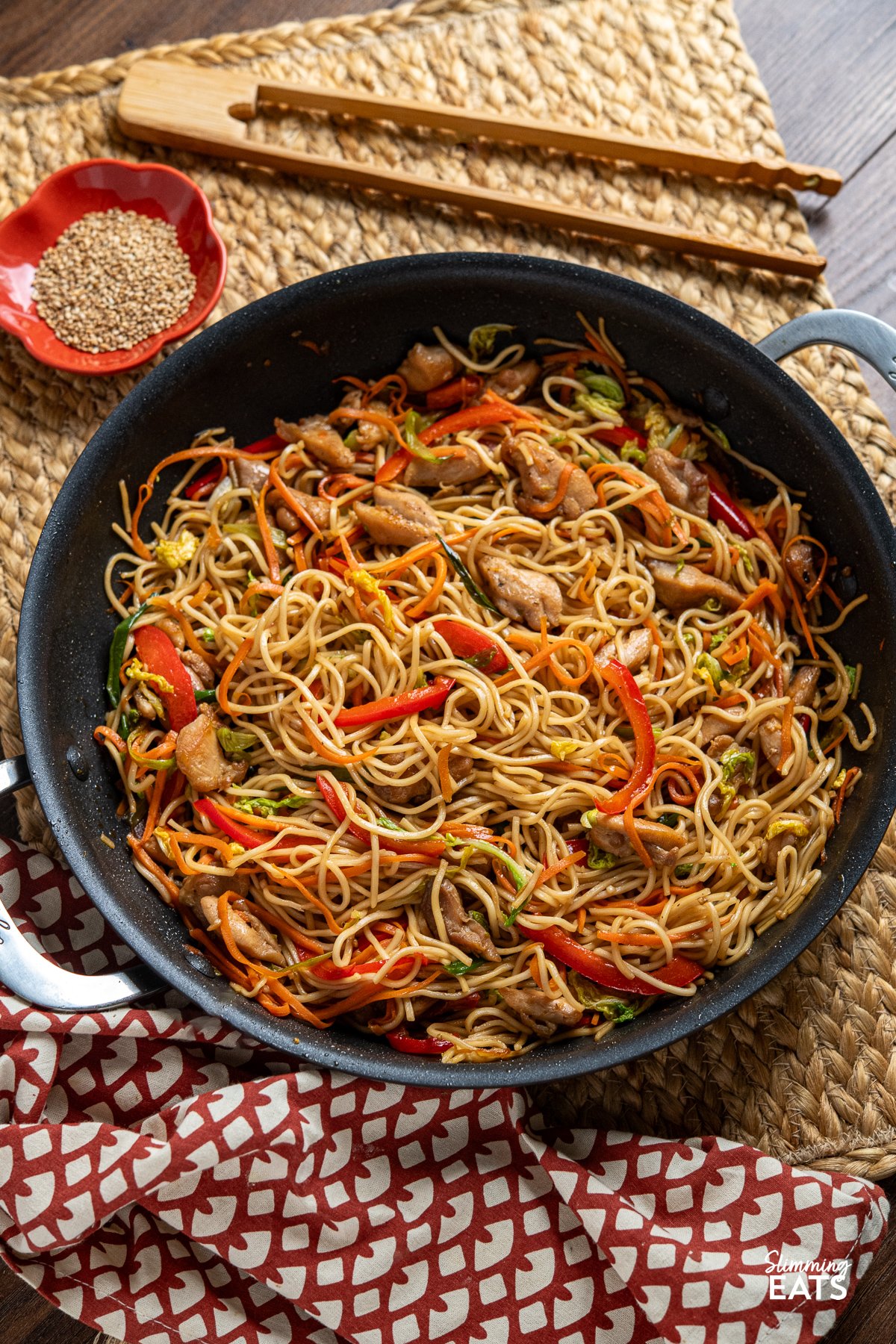 Yakitori Chicken Noodles with Colorful Veggies in a Pan, Accompanied by a Bowl of Toasted Sesame Seeds and Wooden Tongs