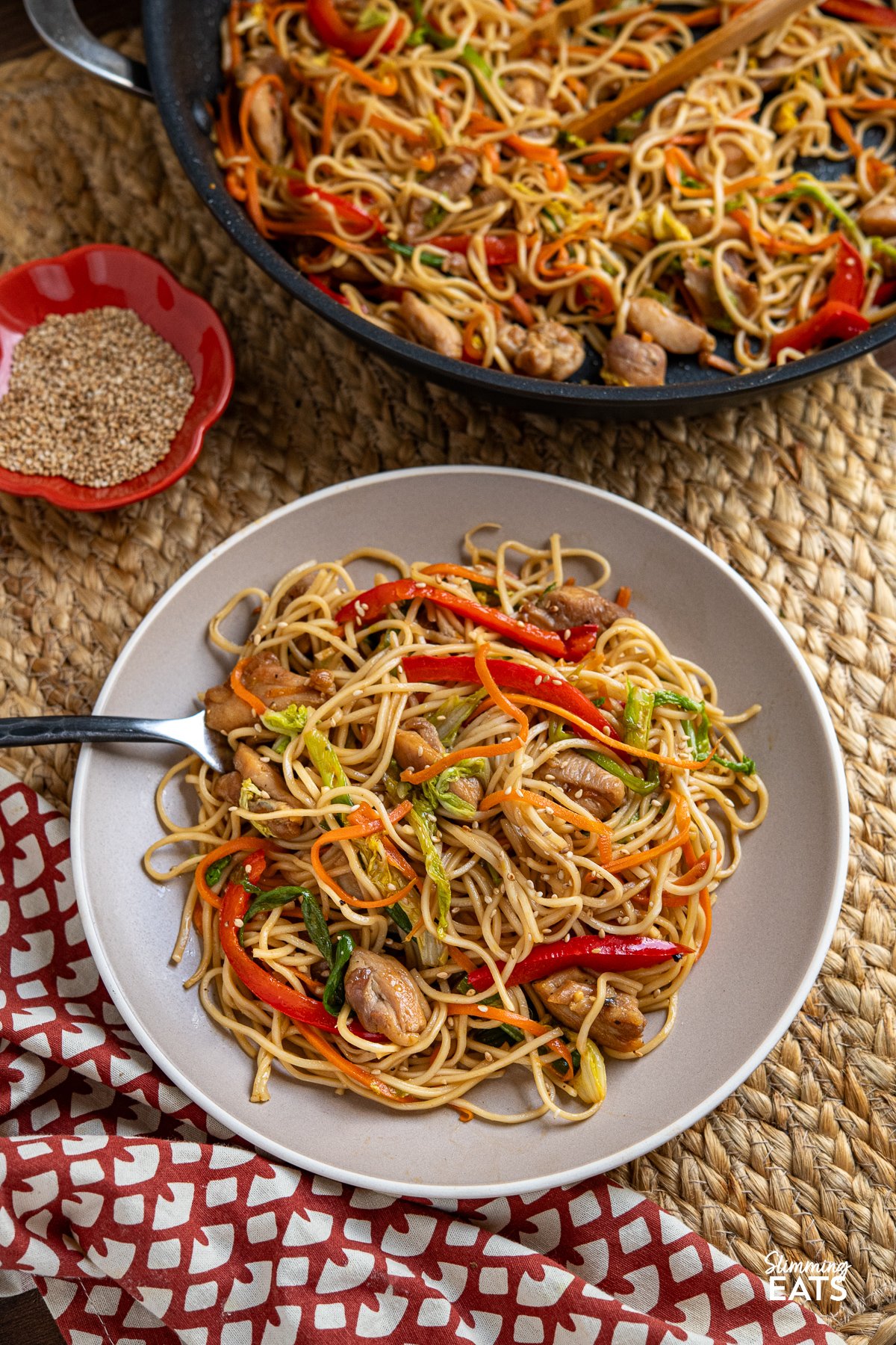 Yakitori Chicken Noodles with Colorful Veggies on a Plate, Fork Resting in Noodles, Pan and Sesame Seeds Bowl in Background