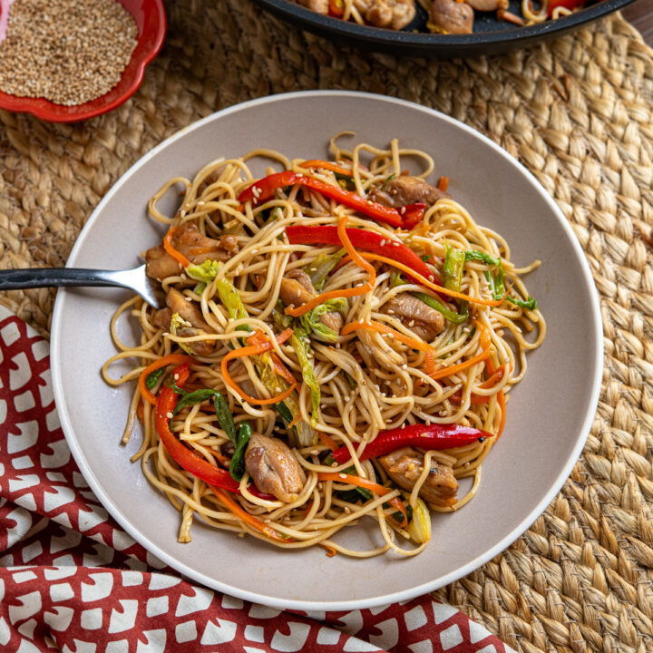 Yakitori Chicken Noodles with Colorful Veggies on a Plate, Fork Resting in Noodles, Pan and Sesame Seeds Bowl in Background