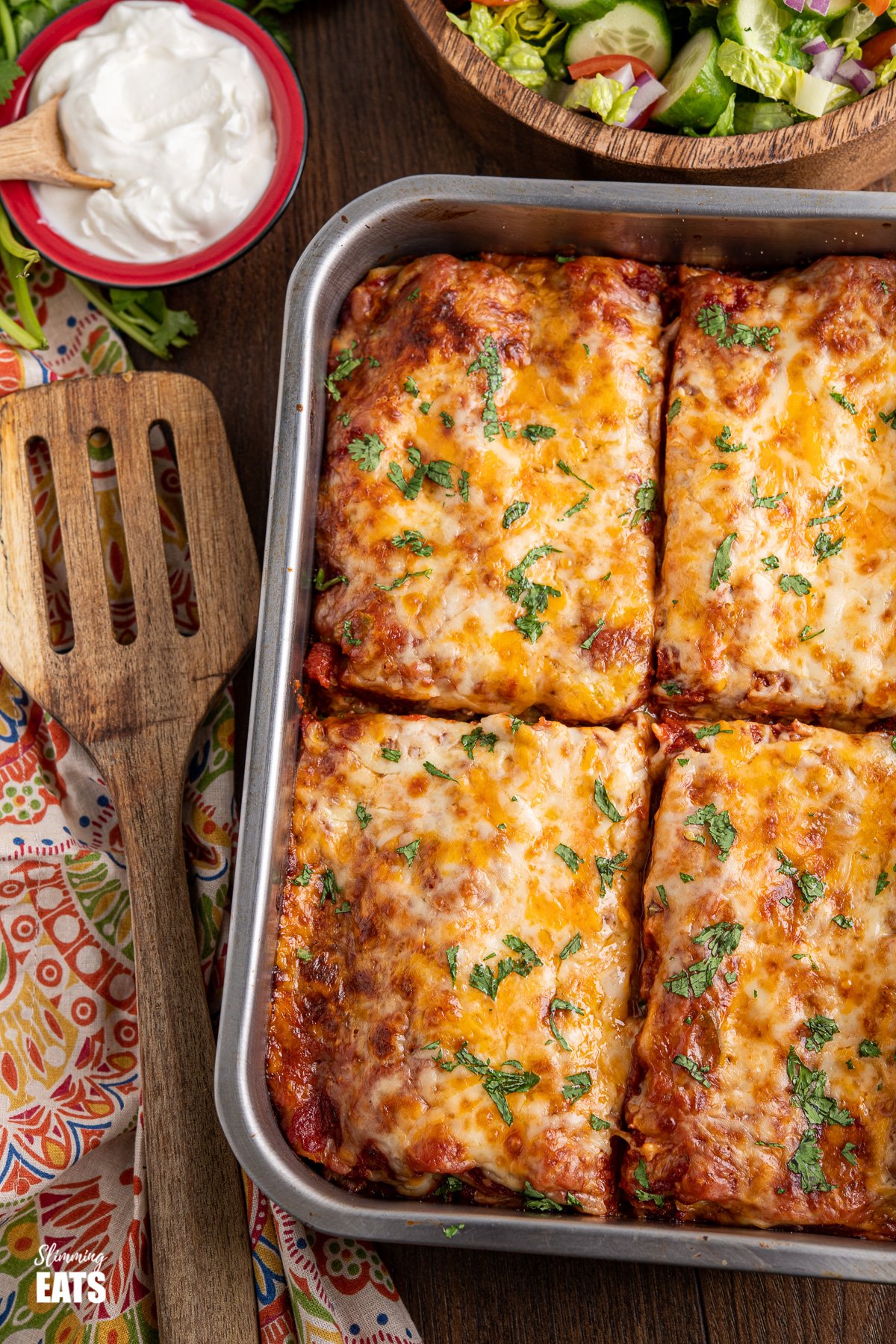 sliced pasta chicken enchiladas in metal baking tray with soured cream and salad in background