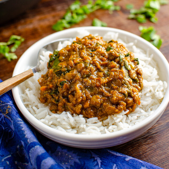 Spicy lentils and spinach served in a white bowl with rice, accompanied by a spoon on the left side. A frying pan and scattered coriander in the background