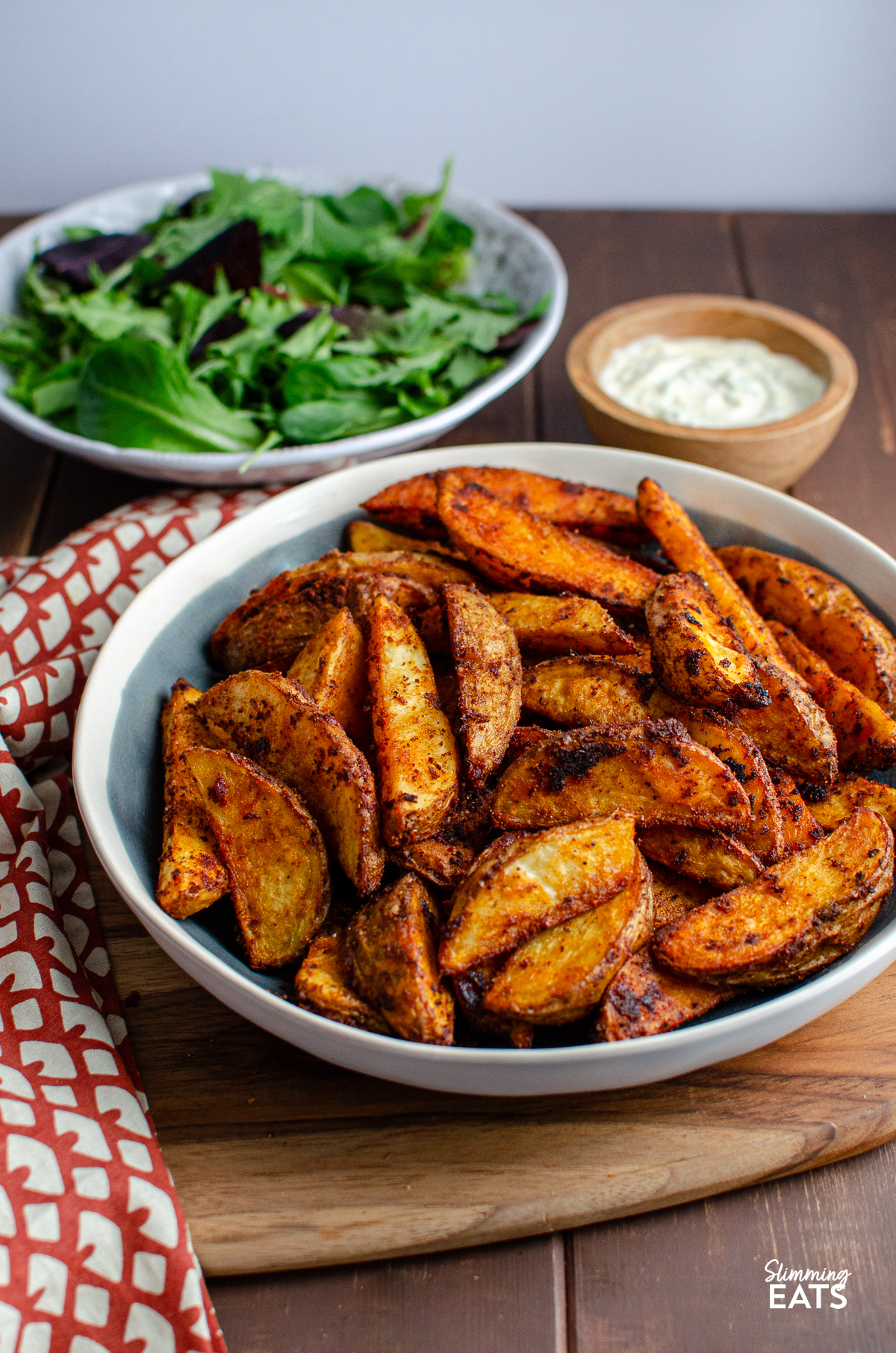 Spicy potato wedges in a blue-gray bowl with white rim, accompanied by salad and garlic dip in the background.