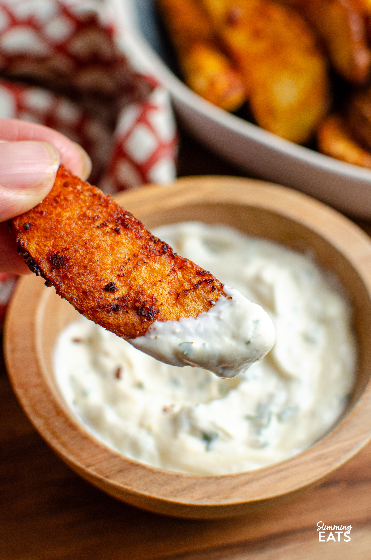 Dipping a spicy potato wedge into garlic dip in a small wooden bowl.