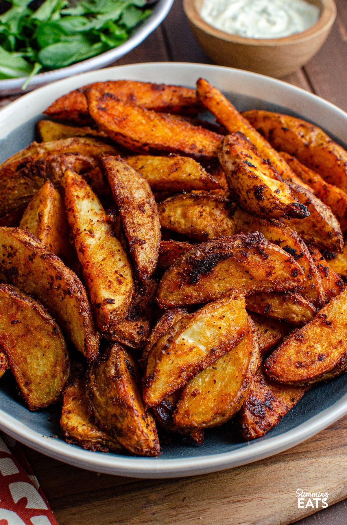 Spicy potato wedges in a blue-gray bowl with white rim, accompanied by salad and garlic dip in the background.