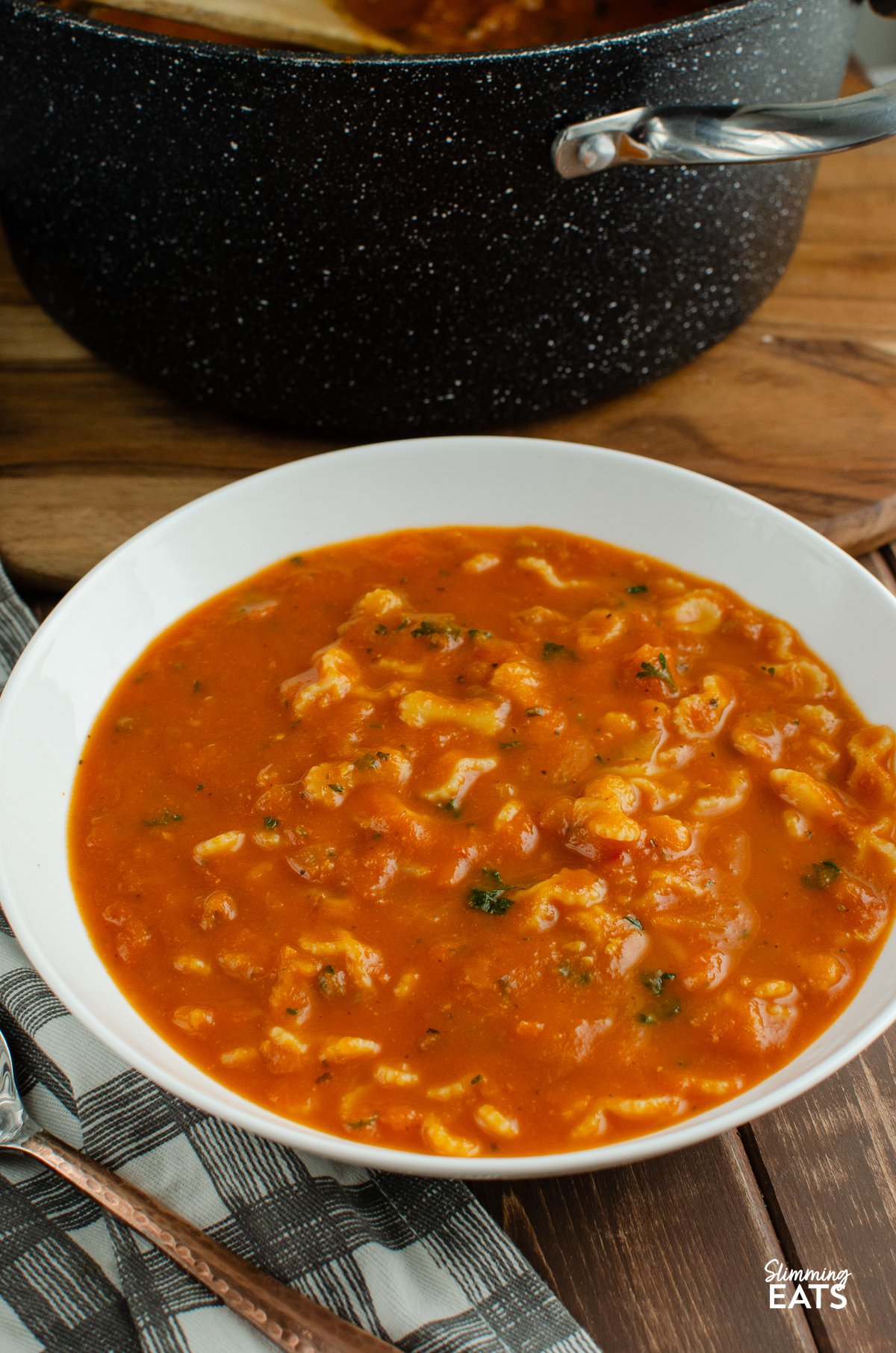 Tomato and pasta soup served in a white bowl accompanied by a spoon with a golden handle, with a saucepan of extra soup visible in the background, suggesting a warm and inviting meal setting.