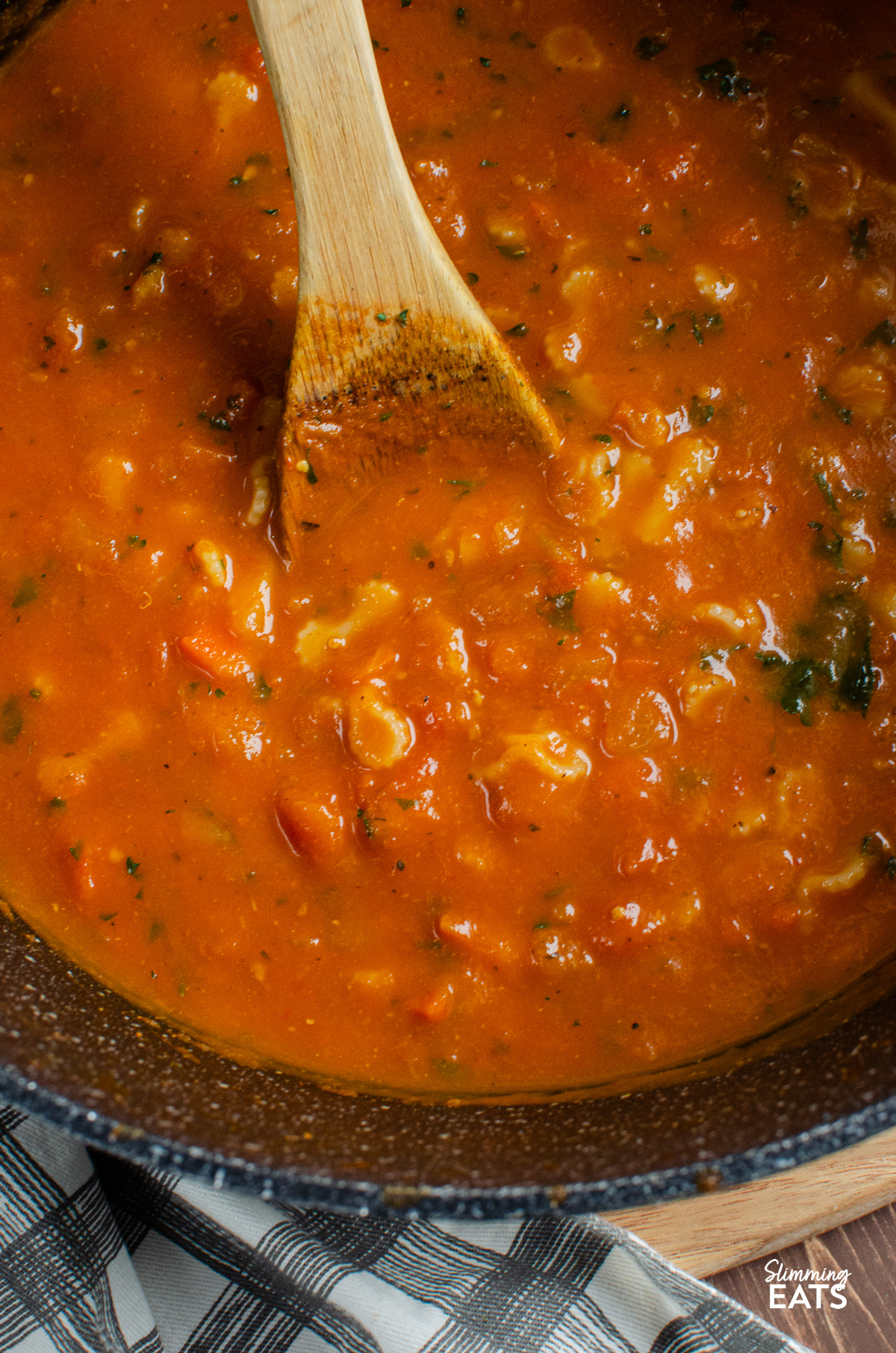 close up of A rich and vibrant tomato and pastasoup simmering in a large black ceramic non-stick saucepan, with a wooden spoon resting inside, ready for stirring.