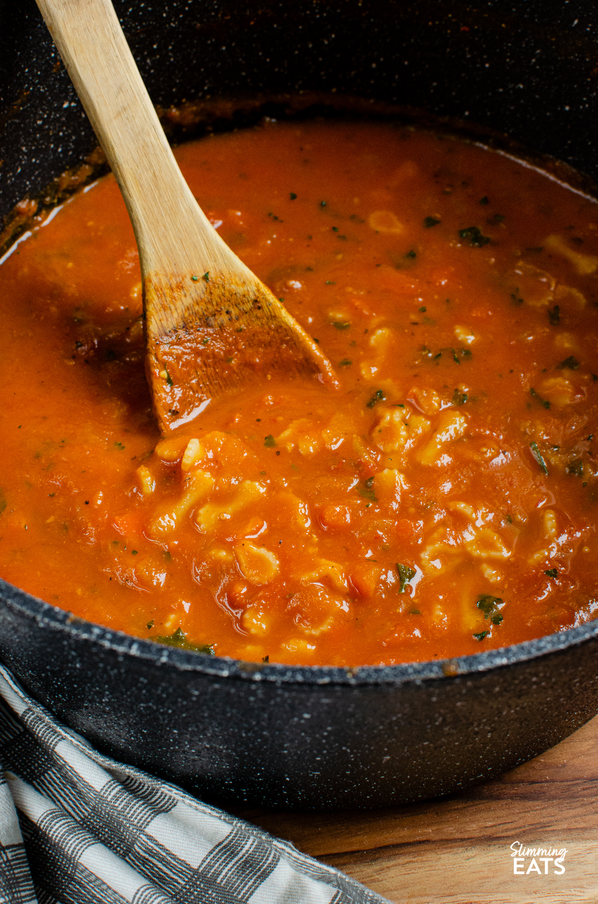 A rich and vibrant tomato and pasta soup simmering in a large black ceramic non-stick saucepan, with a wooden spoon resting inside, ready for stirring.