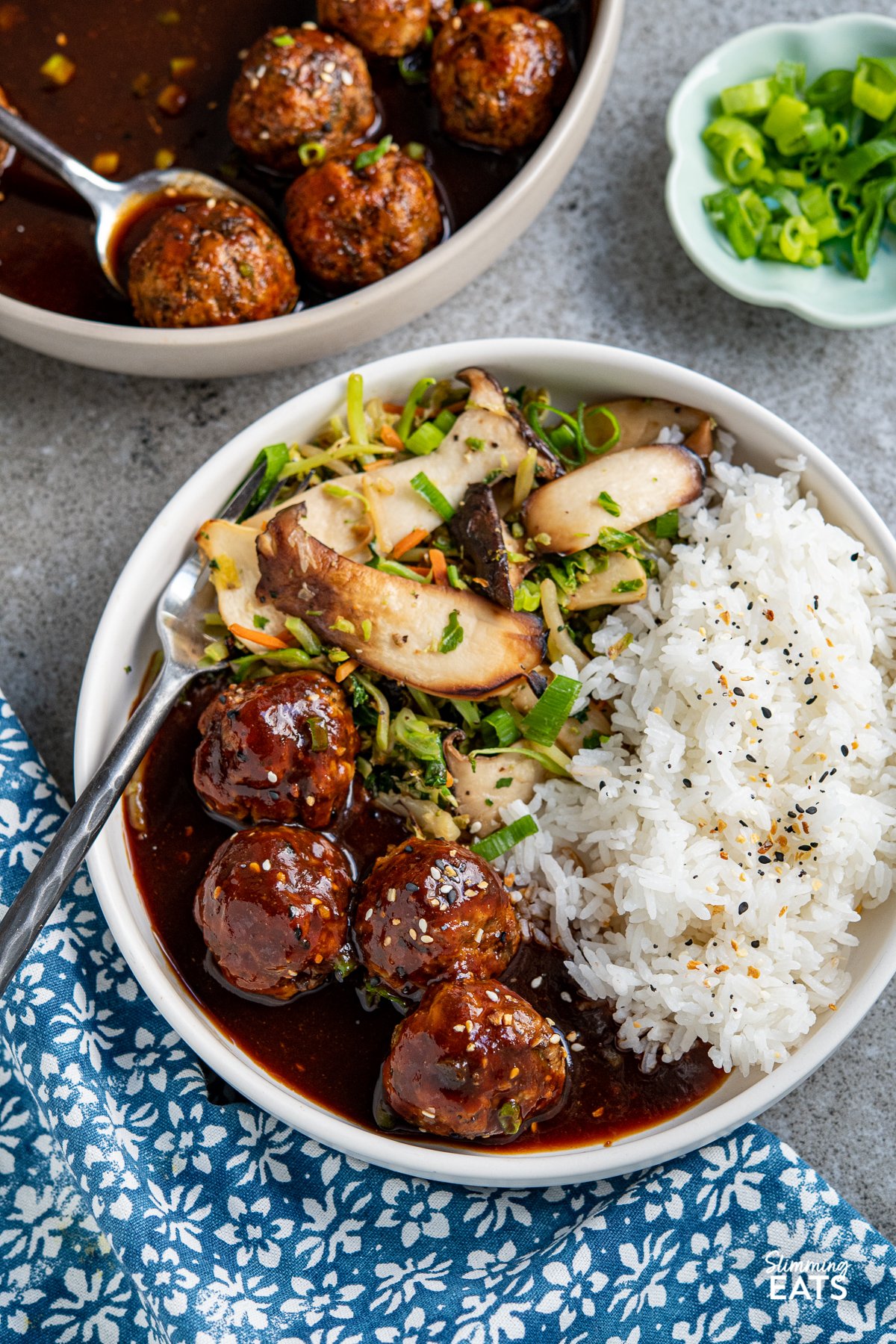 serving of Chicken Meatballs in white bowl with jasmine rice and stir fried vegetables, bowl of meatballs and small blue bowl of chopped spring onions in background