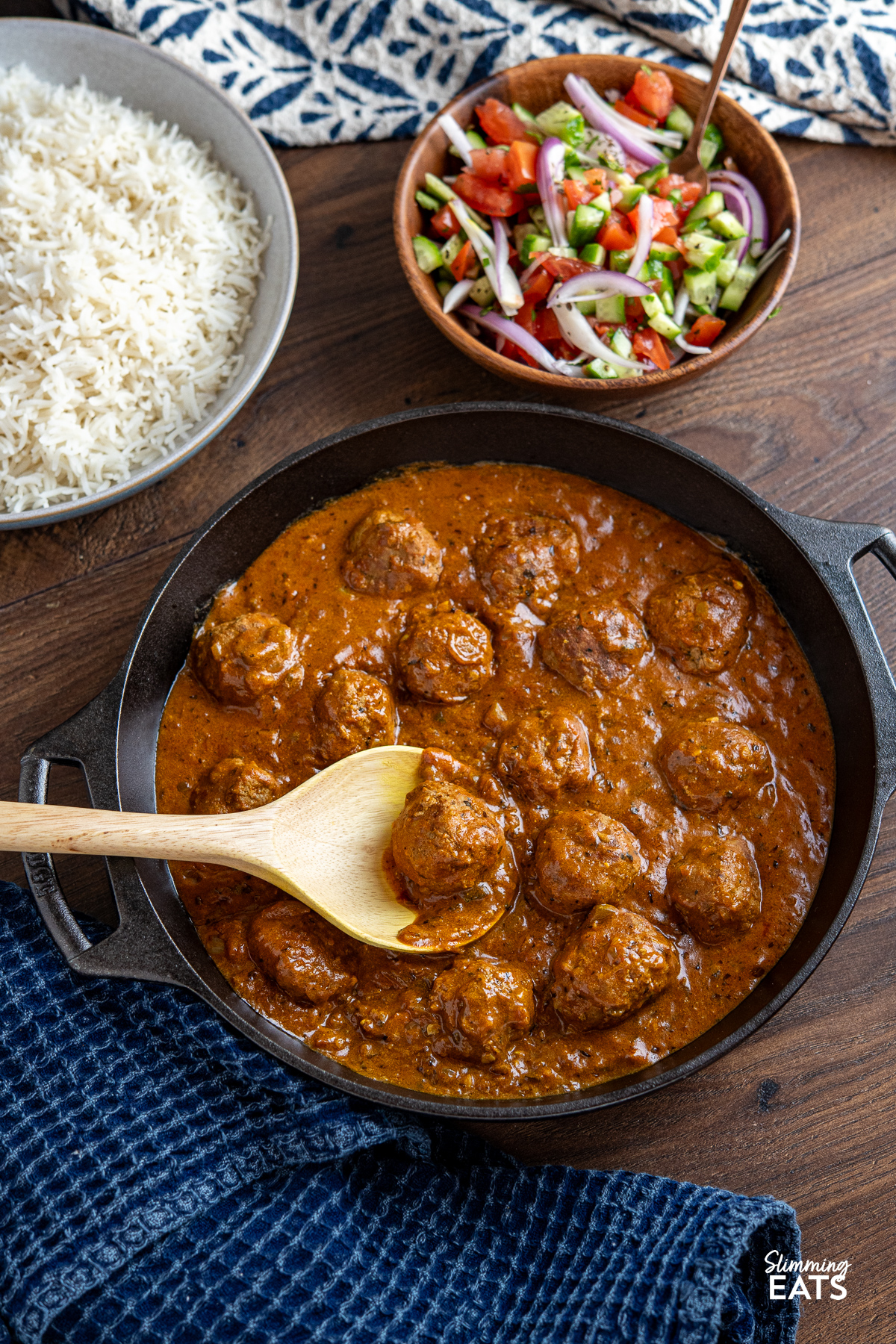 A cast iron double-handled skillet containing lamb kofta curry, placed in the foreground. In the background, there is a bowl of rice and a bowl of vibrant Indian salad, featuring fresh vegetables and herbs.