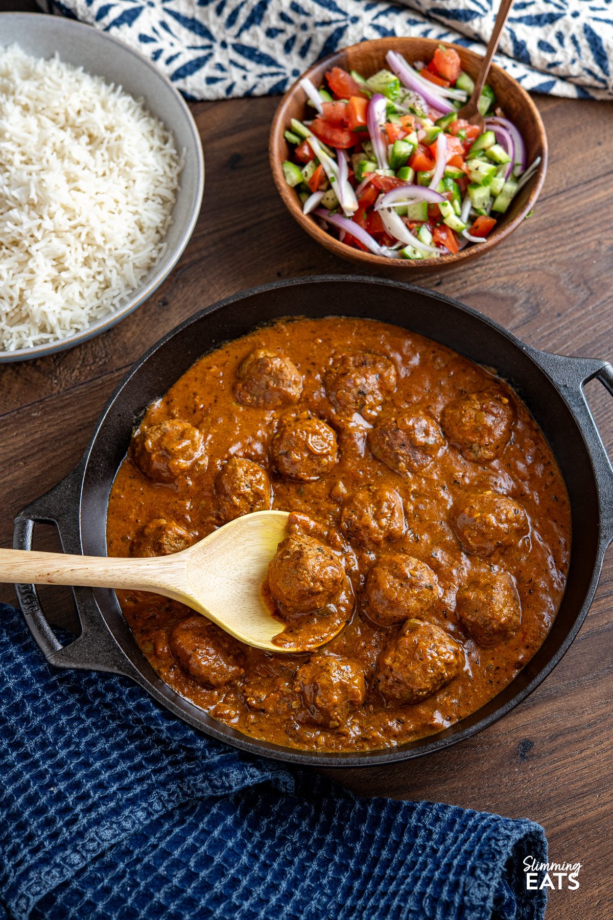 A cast iron double-handled skillet containing lamb kofta curry, placed in the foreground. In the background, there is a bowl of rice and a bowl of vibrant Indian salad, featuring fresh vegetables and herbs.