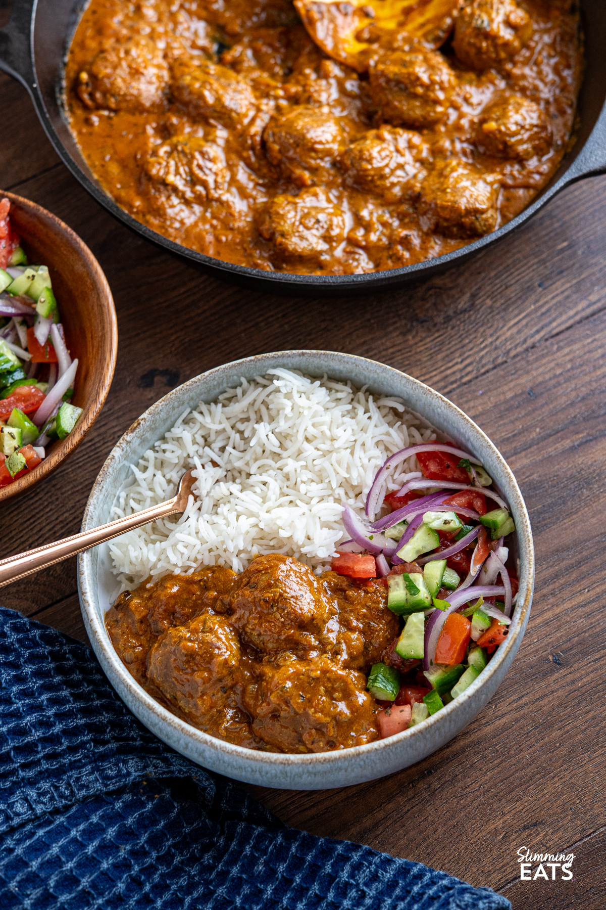 
A pale blue bowl filled with lamb kofta curry and rice, accompanied by a fork, with a skillet of additional kofta and a bowl of colorful Indian salad in the background.