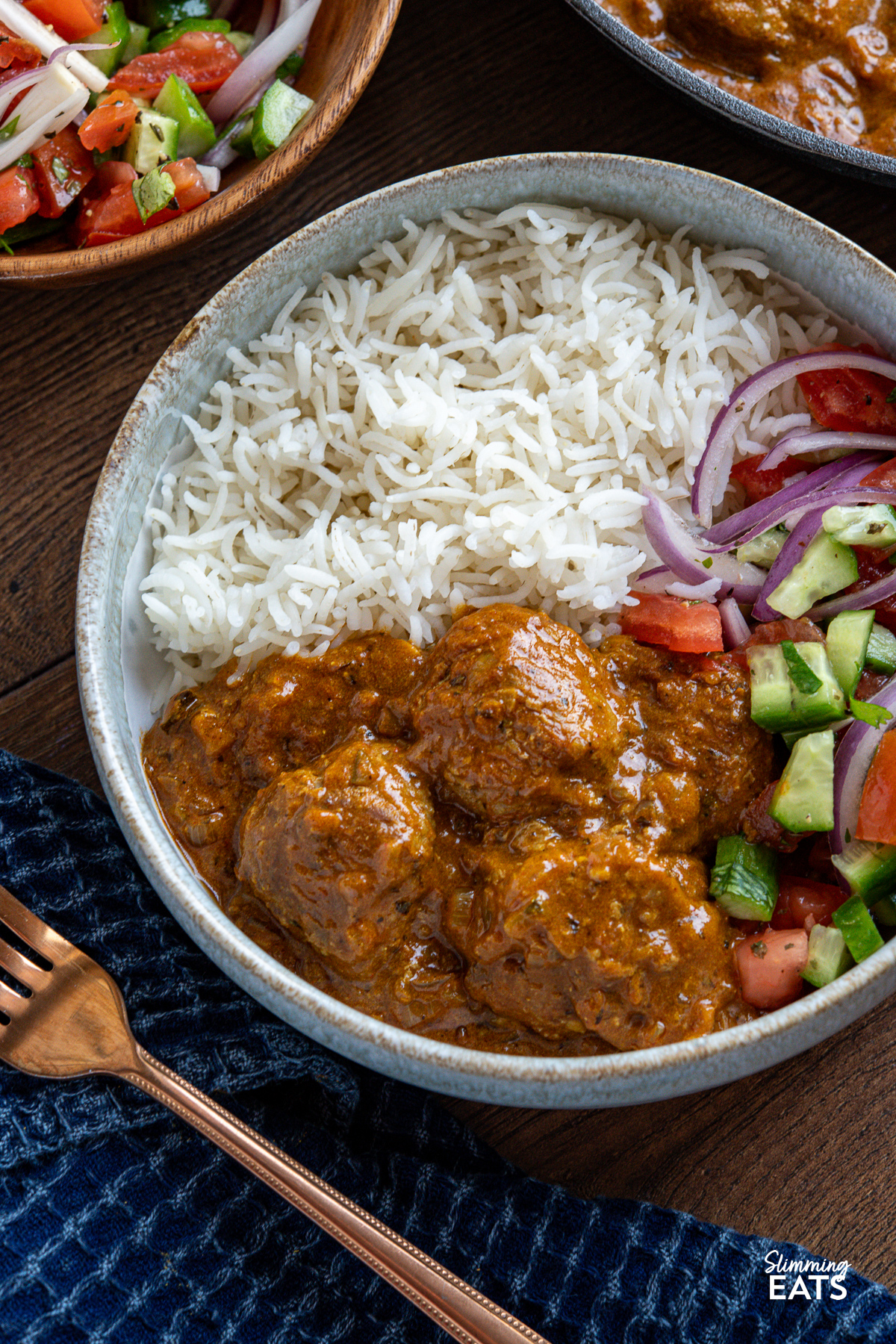 close up of 
A pale blue bowl filled with lamb kofta curry and rice, accompanied by a fork, with a skillet of additional kofta and a bowl of colorful Indian salad in the background.
