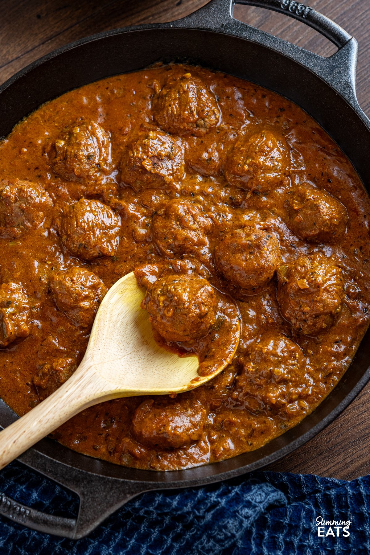 A cast iron double-handled skillet containing lamb kofta curry with a wooden spoon placed in pan