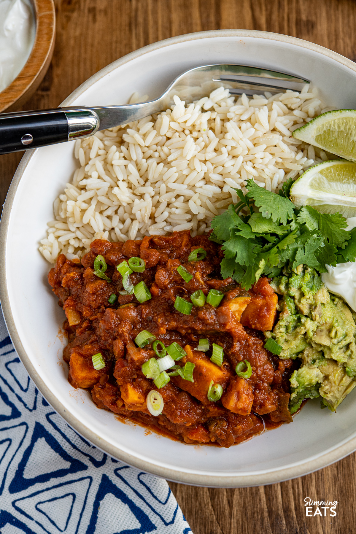close up of Sweet Potato, Vegetable and Lentil Chilli in a white bowl with blue stripes,   served with cheddar, brown rice, lime wedges, sour cream, cilantro and mashed avocado 