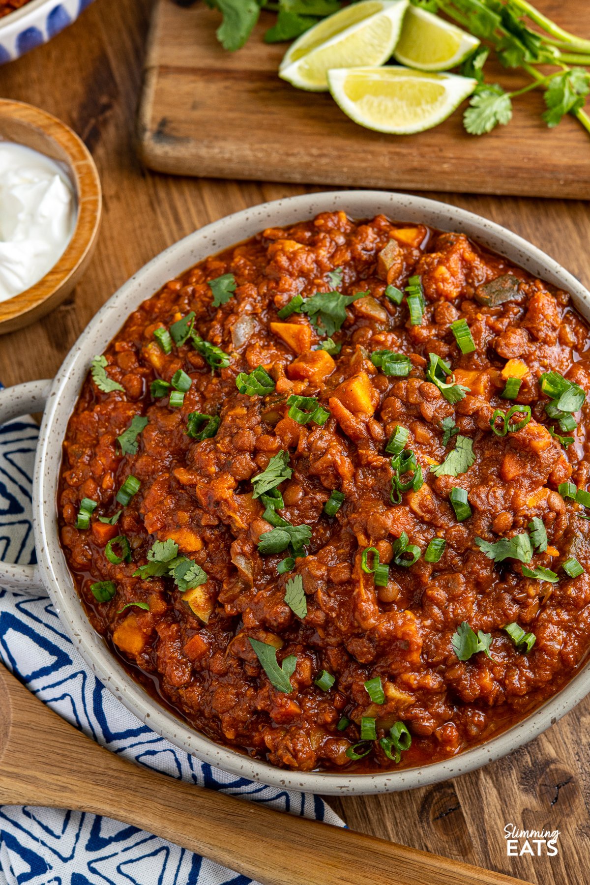 Sweet Potato, Vegetable and Lentil Chilli in a brown grey double handled dish with sour cream, lime wedges and cilantro in background