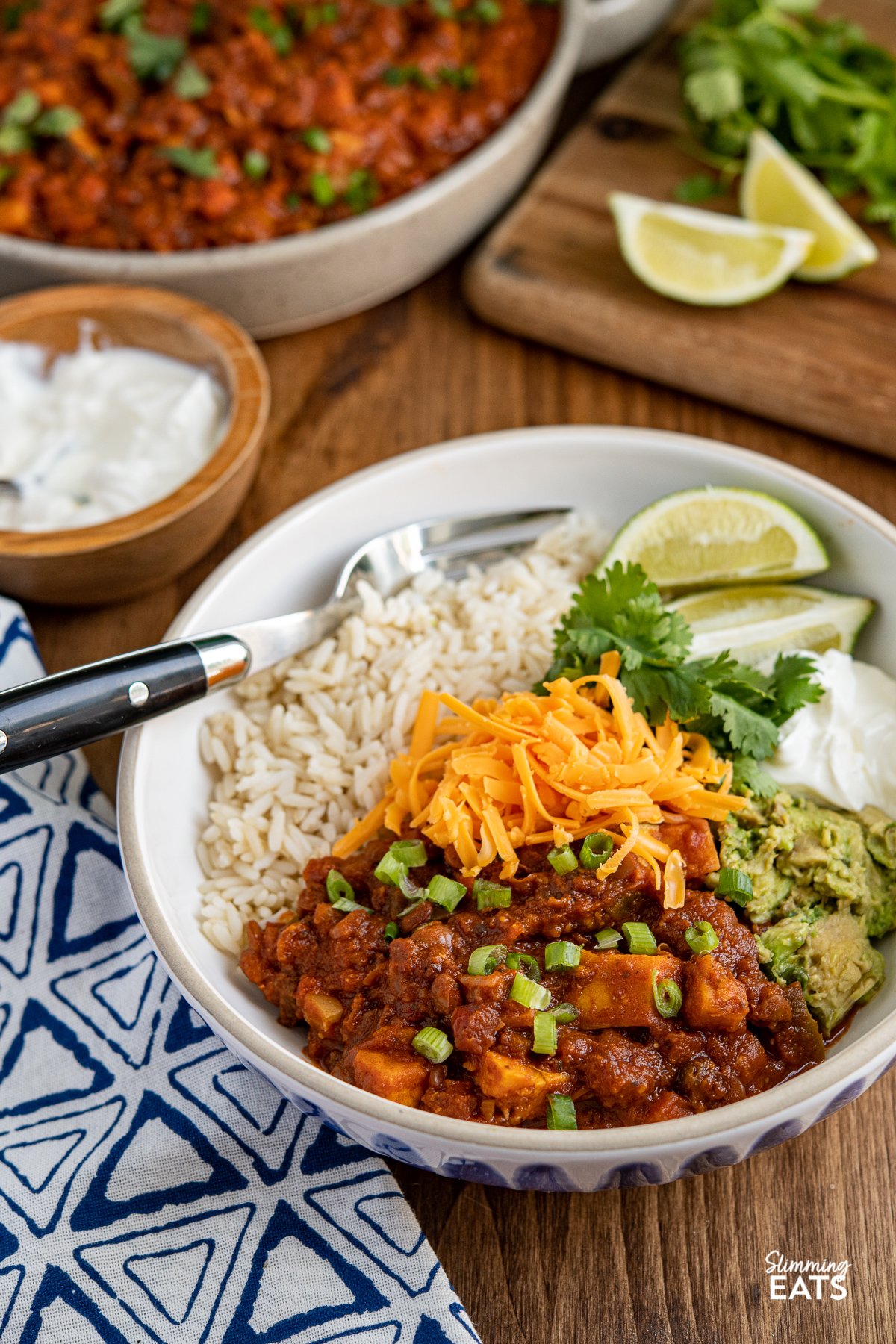 Sweet Potato, Vegetable and Lentil Chilli in a white bowl with blue stripes,   served with cheddar, brown rice, lime wedges, sour cream, cilantro and mashed avocado 