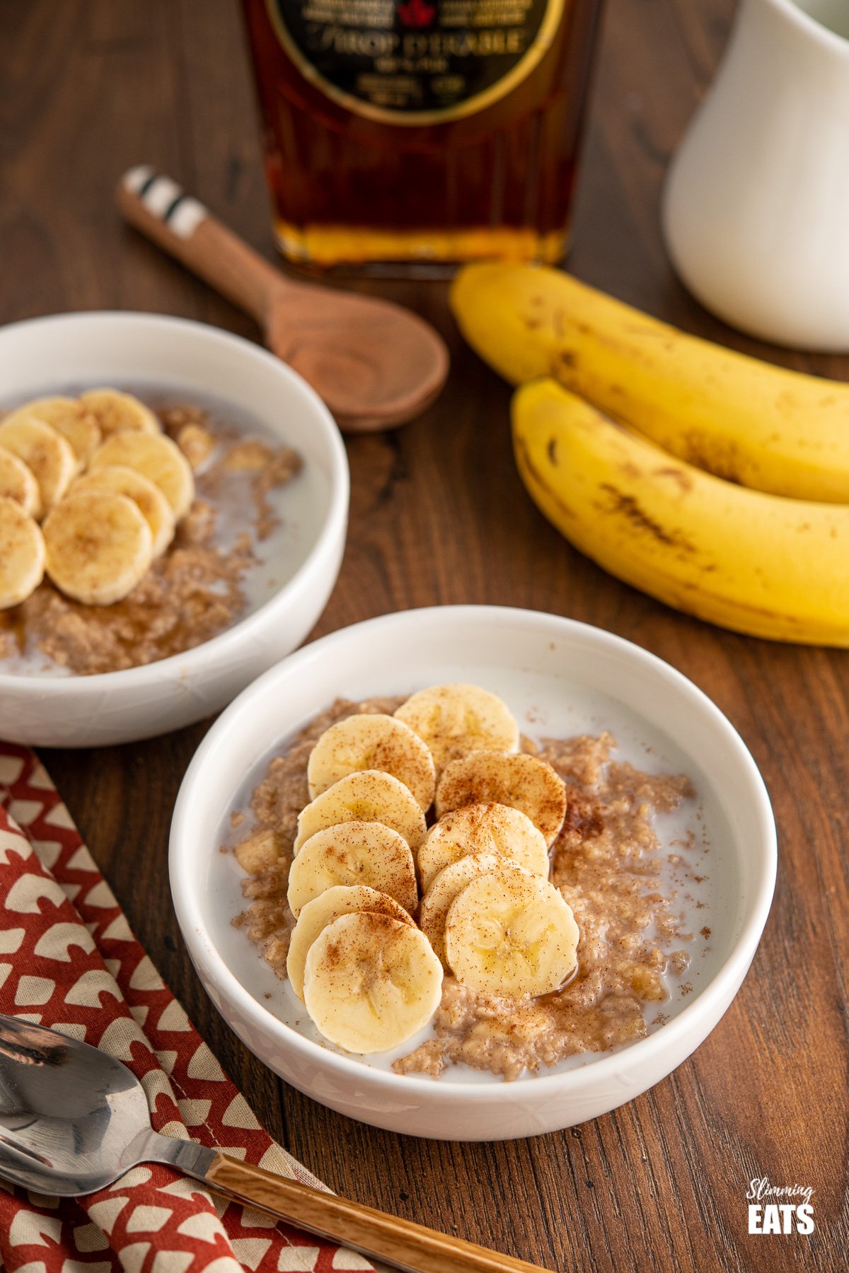 two bowls of creamy milky oatmeal topped with slices of banana, cinnamon and a drizzle of maple syrup, two banans and maple syrup bottle in the background