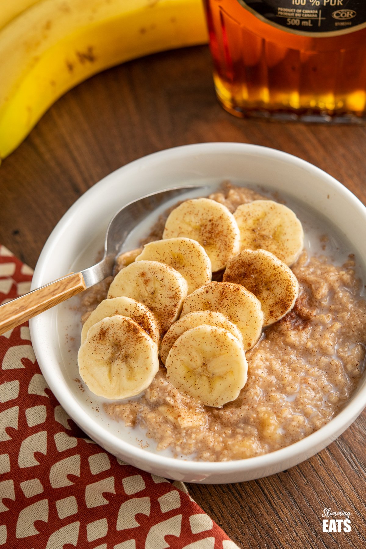 bowl of creamy milky banana cinnamon maple oatmeal with wooden handled spoon and banana and maple syrup bottle in the background