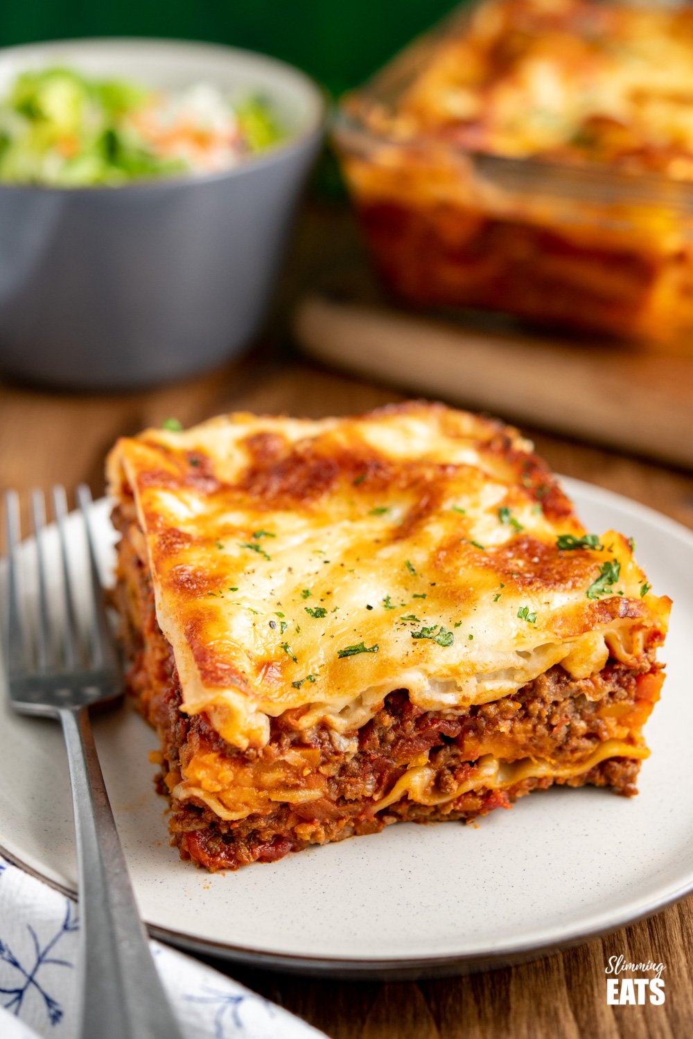 The BEST Low Syn Beef Lasagne - amazingly rich beef sauce in between layers of pasta and sweet potato, all finished off with a delicious healthier bechamel white sauce and melted cheese. beef lasagne on cream plate with silver fork - salad and lasagne in background.