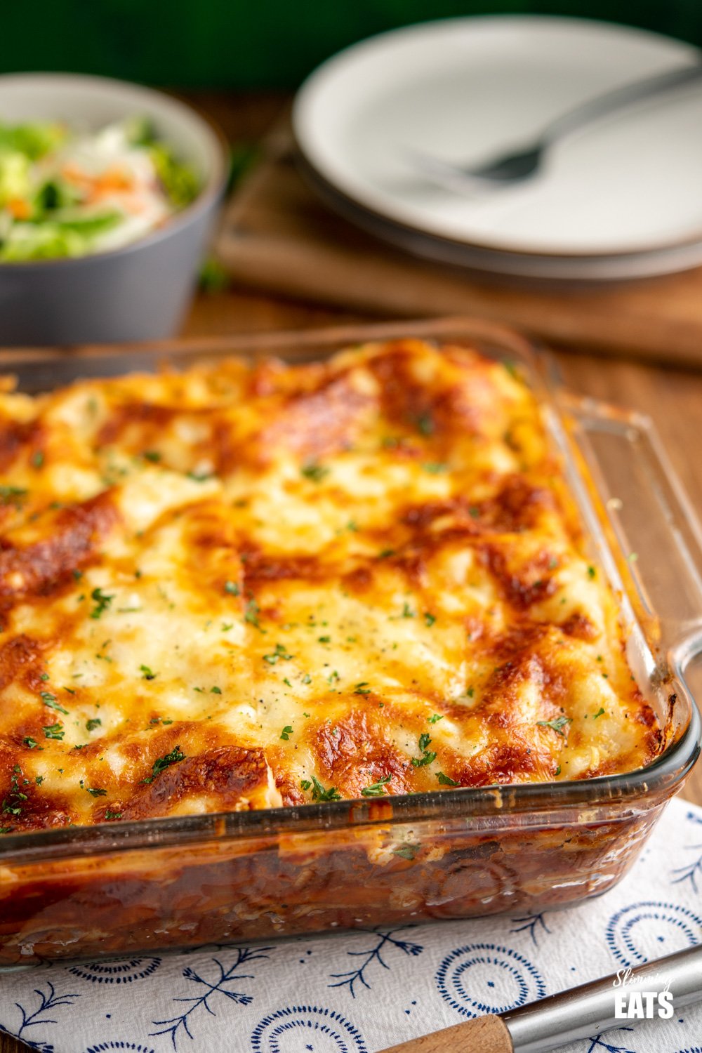 Beef Lasagne in pyrex dish on white and blue placemat with plates and salad bowl in background 