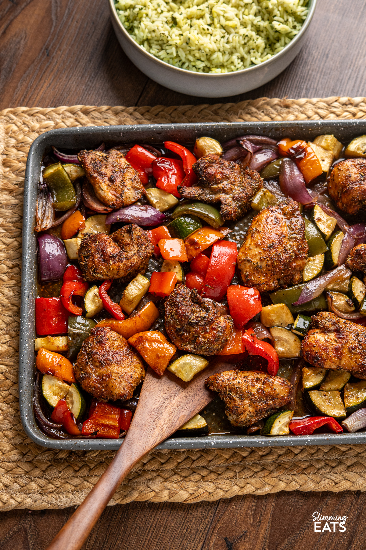 Black pepper chicken with balsamic roasted vegetables arranged on a baking tray, accompanied by a bowl of herby seasoned rice in the background