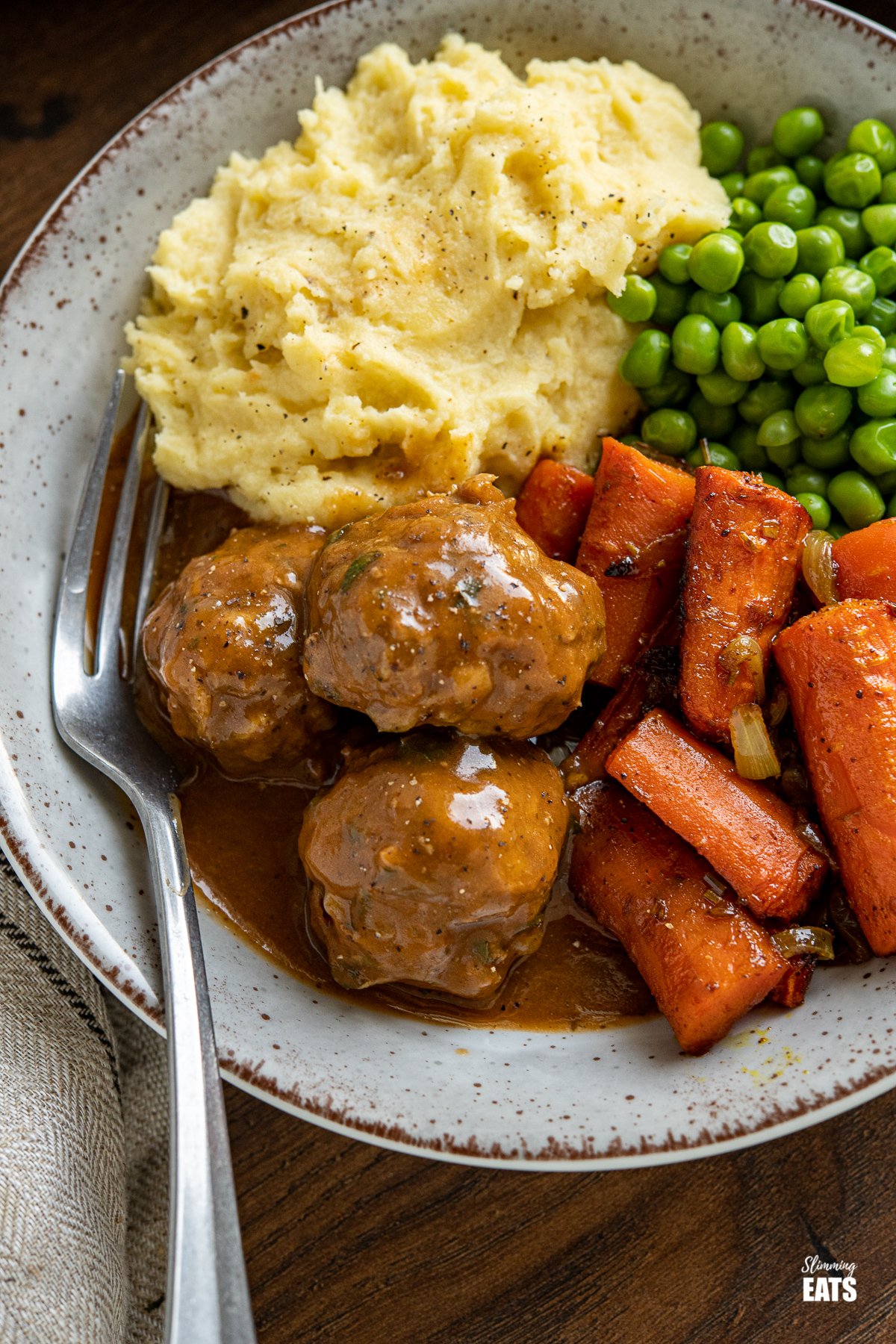 close up of Lamb Meatballs with Mint Gravy in a grey and brown bowl with carrots, onion, peas and mashed potatoes