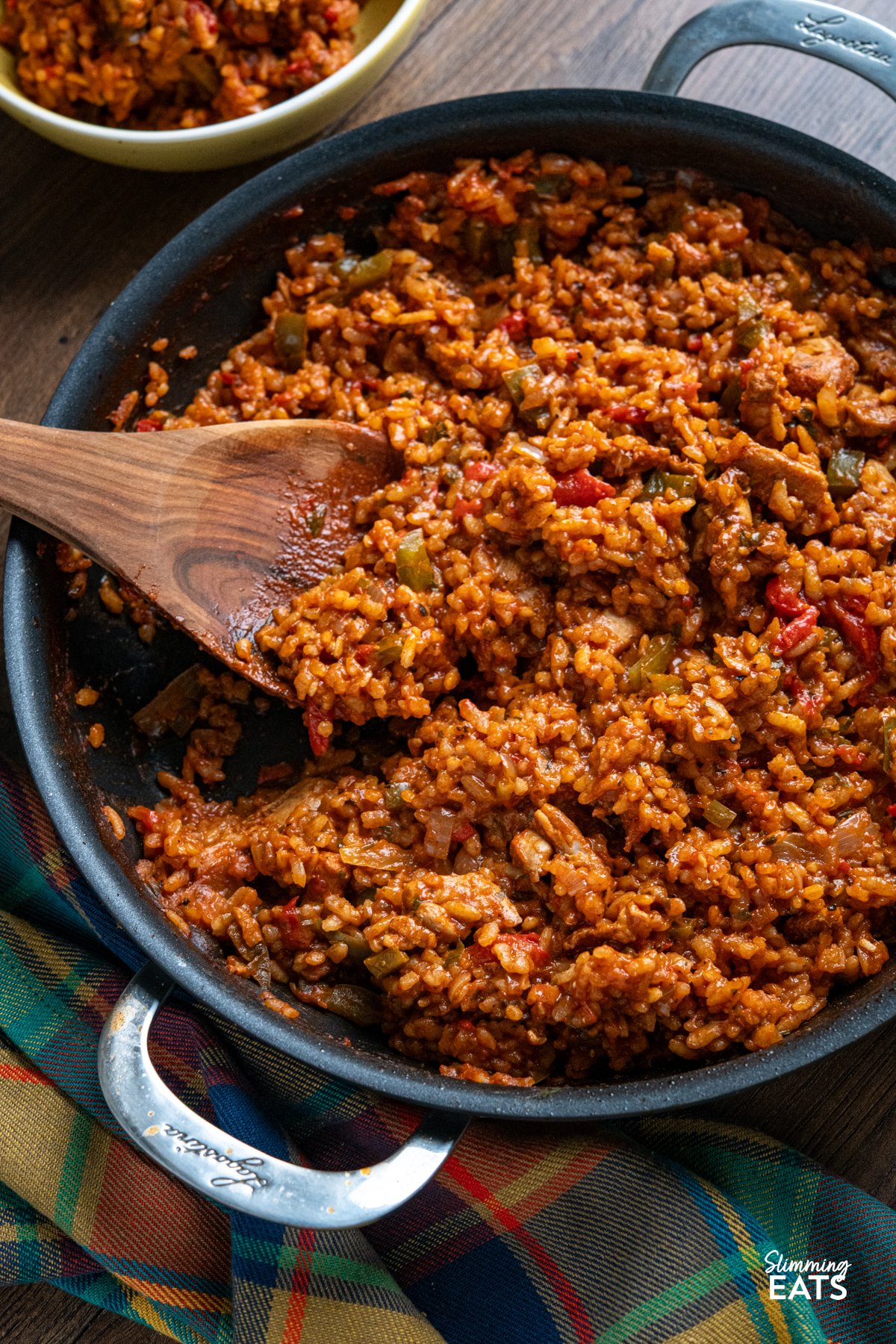 One Pot Spicy Spanish Chicken with Rice – a bold, risotto-style dish with tender chicken, smoky paprika, and creamy rice, all cooked in one pan for maximum flavour with minimal effort. Spicy Spanish Chicken Rice in double handled non stick pan with wooden spoon - serving in background in small yellow bowl