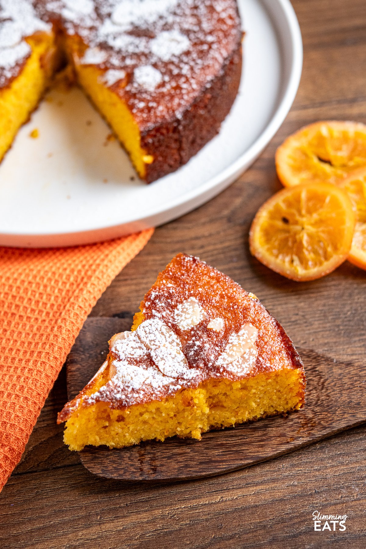 This Flourless Orange Almond Cake is a delightful fusion of almond richness and the zingy essence of oranges. With just a handful of simple ingredients, this grain-free and gluten-free creation yields a moist crumb with a subtle sweetness, perfectly complemented by a burst of vibrant orange flavour. slice of Clementine Orange Almond Cake on a wooden spatula