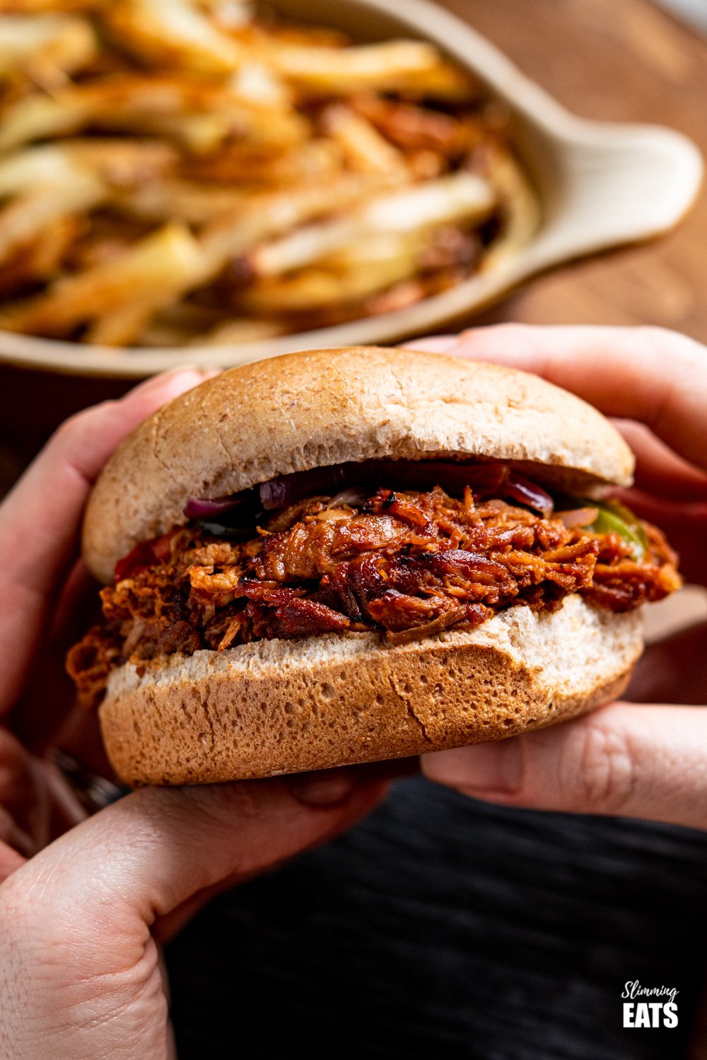 slow cooker pulled pork sandwich held in two hands with crispy oven baked chips in background