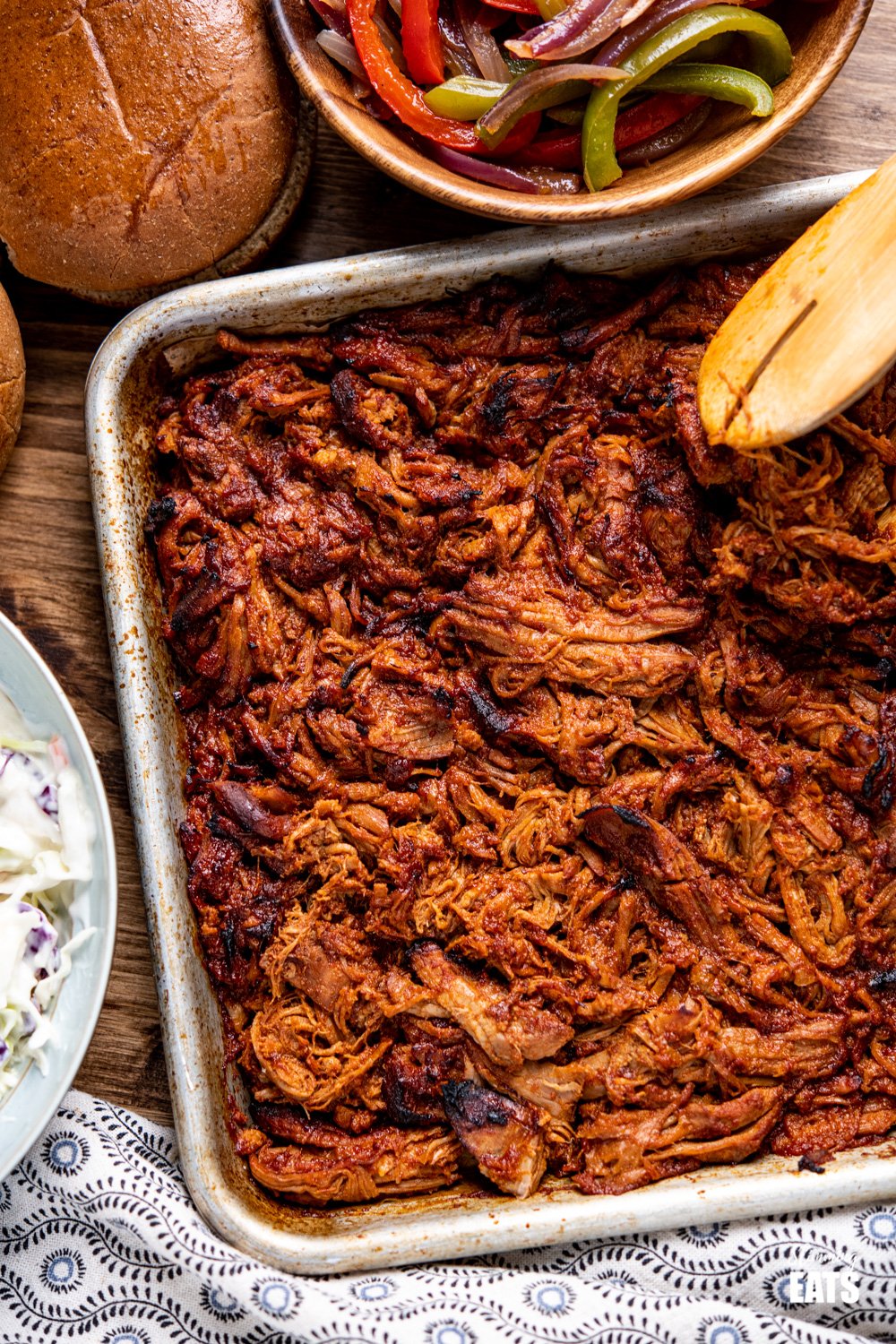 slow cooker pulled pork caramezlied on baking tray with a bowl of peppers and onions and whole meal rolls.