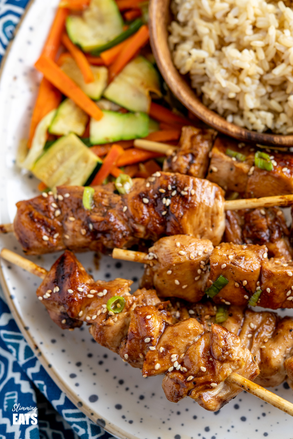 close up Yakitori Chicken Skewers on speckled plate with a bamboo bowl of brown rice and side fo sesame vegetables